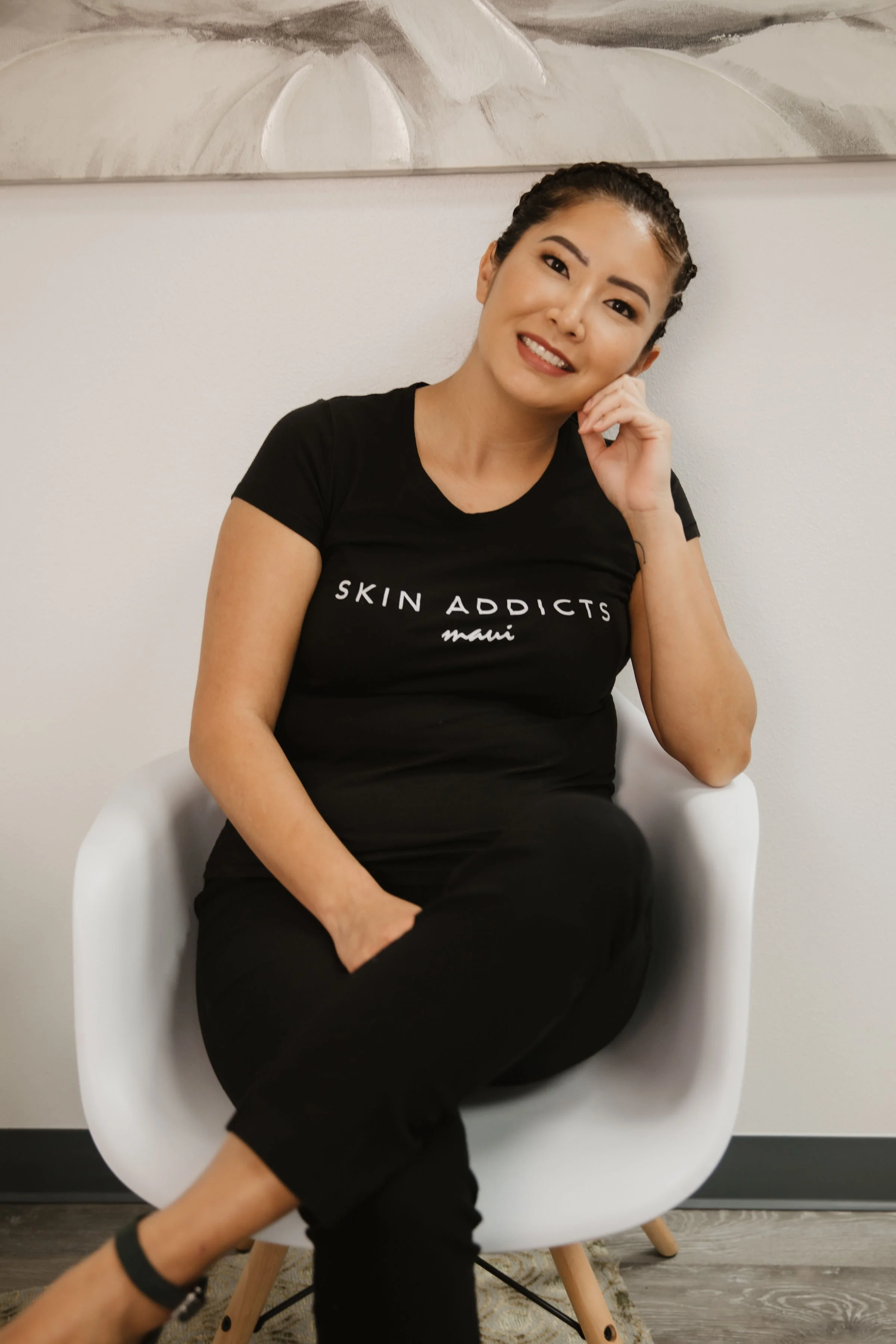 A woman sitting in a white modern chair, smiling and touching her face, wearing a black t-shirt that says 'Skin Addicts Maui' in white and black pants.