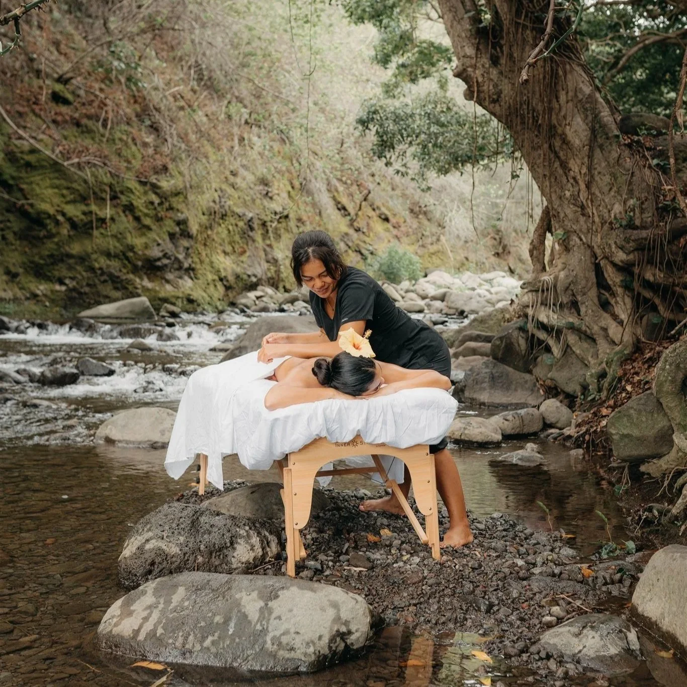 A woman receives a massage outdoors by a woman in a black shirt, on a massage table set in a stream surrounded by rocks and trees.