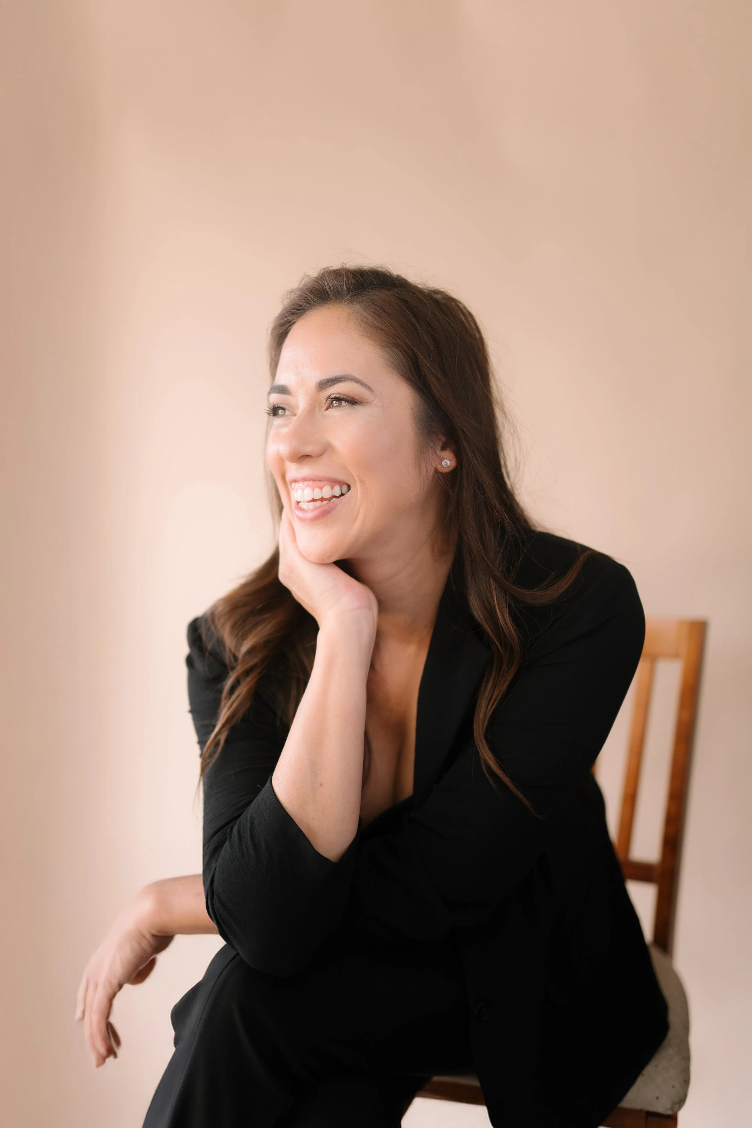A woman with long brown hair, wearing a black blazer, sitting on a wooden chair, smiling and posing with her hand resting on her face against a neutral background.