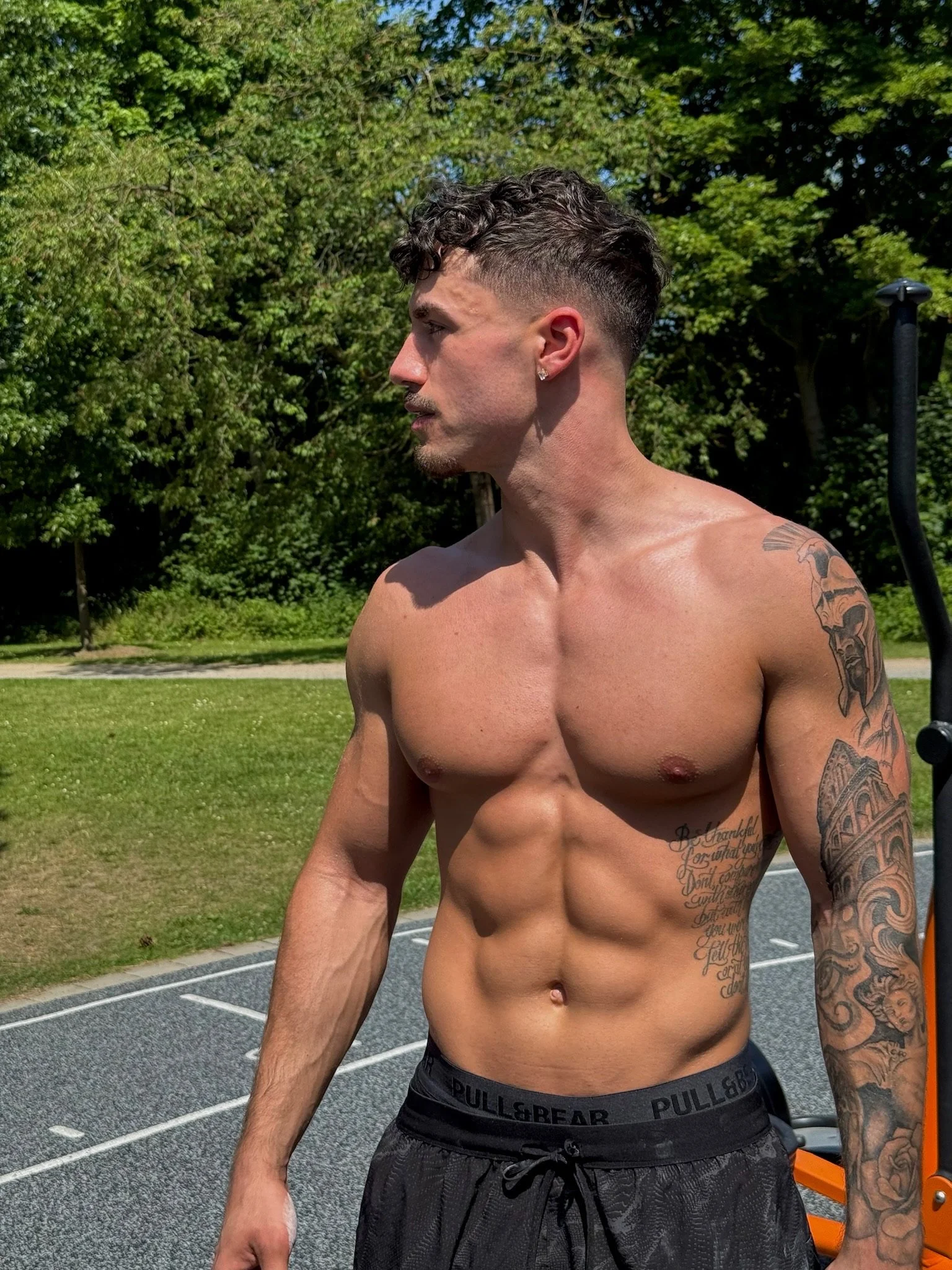 A shirtless man with short, curly dark hair, earrings, and tattoos on his left arm and torso, standing outdoors on a sunny day near some workout equipment with a sports court and green trees in the background.