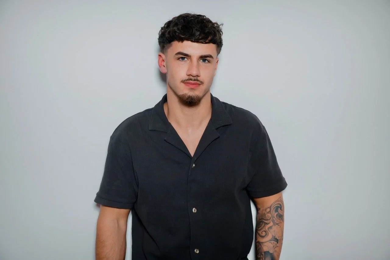 A young man with short, curly dark hair, a goatee, wearing a black short-sleeved button-up shirt, standing against a plain light gray background.