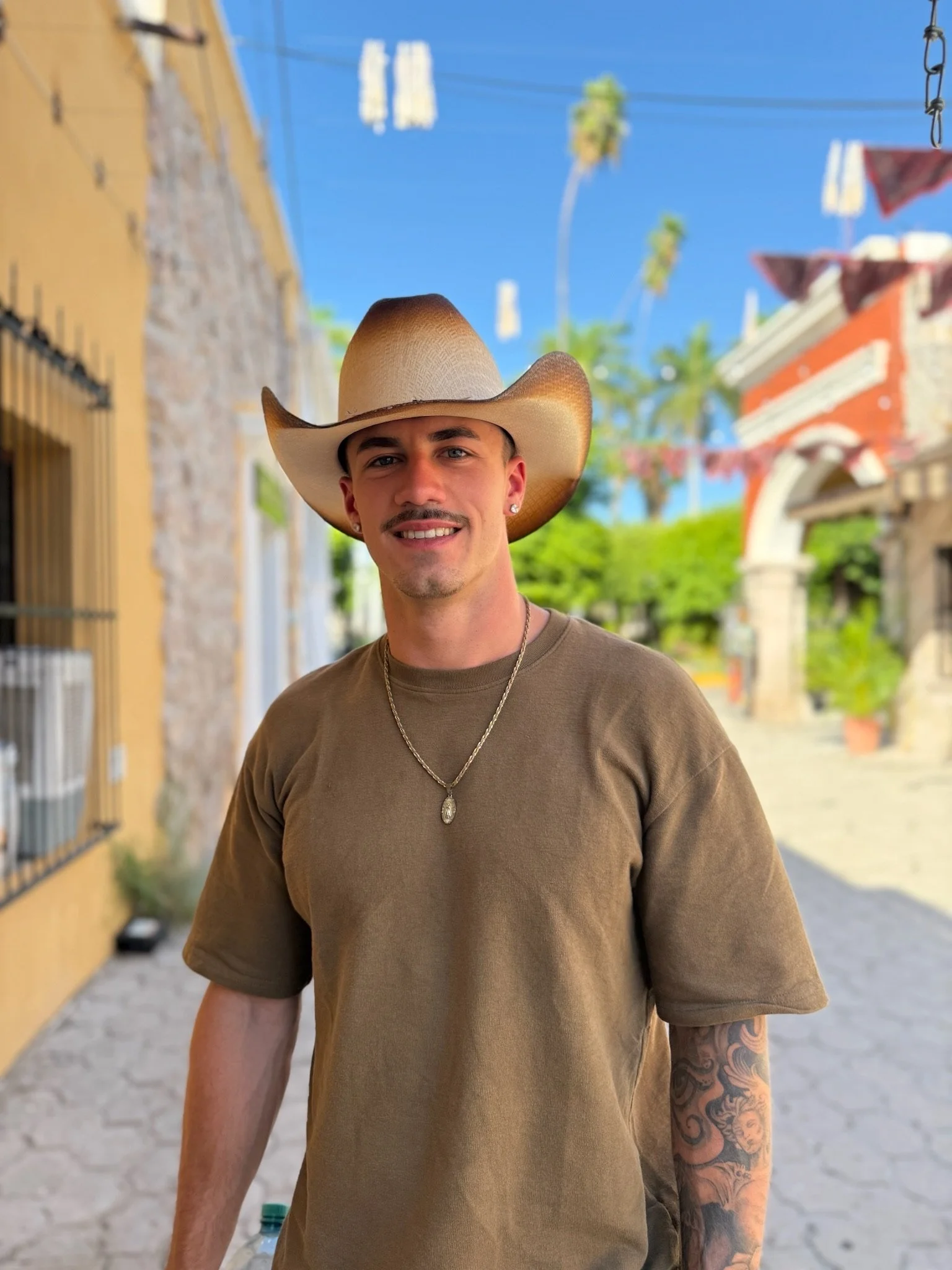 A young man wearing a cowboy hat, tan T-shirt, and jewelry, standing outdoors on a sunny day with a colorful street and palm trees in the background.