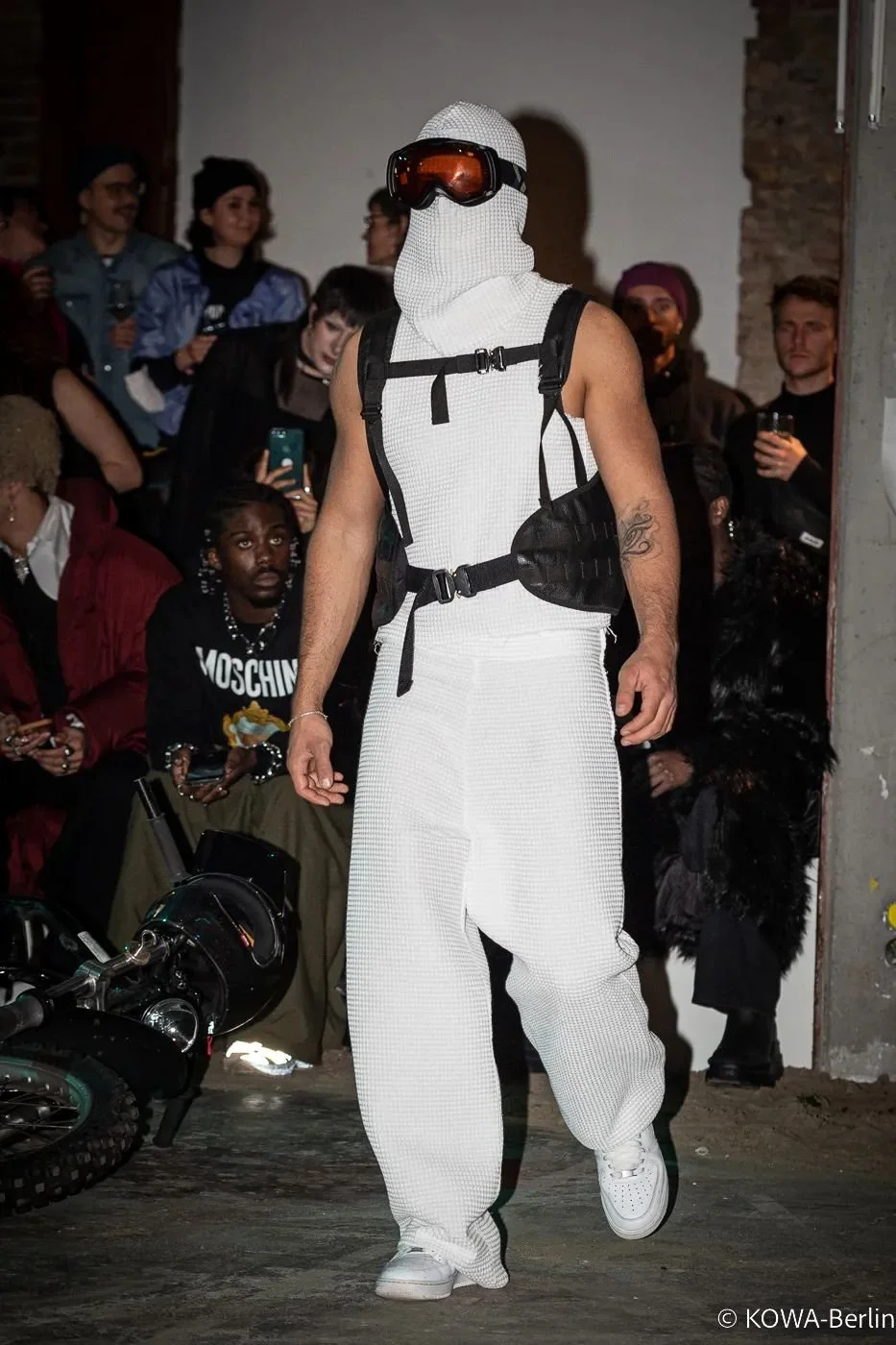 A person dressed in a white scope with a hood and sunglasses with orange lenses walking past a crowd of people at a fashion show or event. The person is wearing white sneakers and carrying black pouches, with a tattoo visible on their left arm. The a