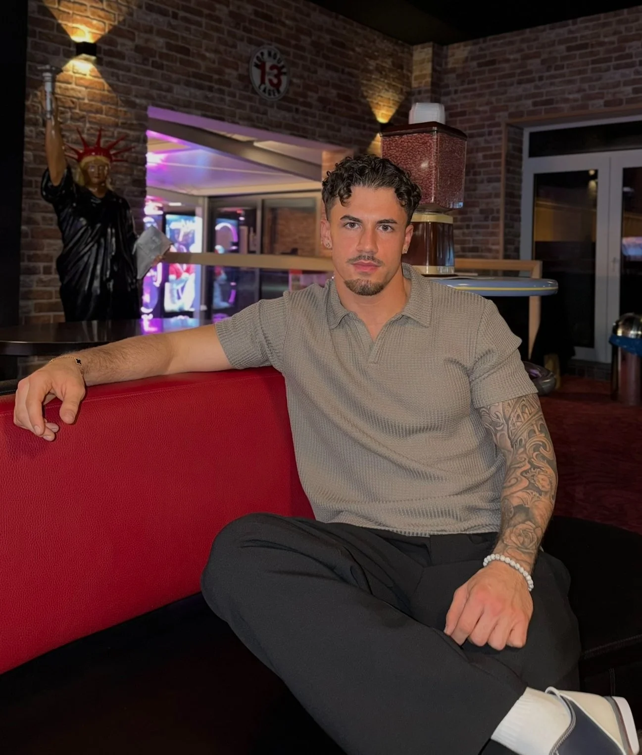 A young man with dark curly hair and a goatee, sitting on a red and black bench inside a bar or restaurant, wearing a beige short-sleeved polo shirt, black pants, and white shoes. He has a tattoo sleeve on his left arm and a white beaded bracelet on his left wrist. In the background, a trophy-style statue of the Statue of Liberty and a brick wall with neon lighting are visible.