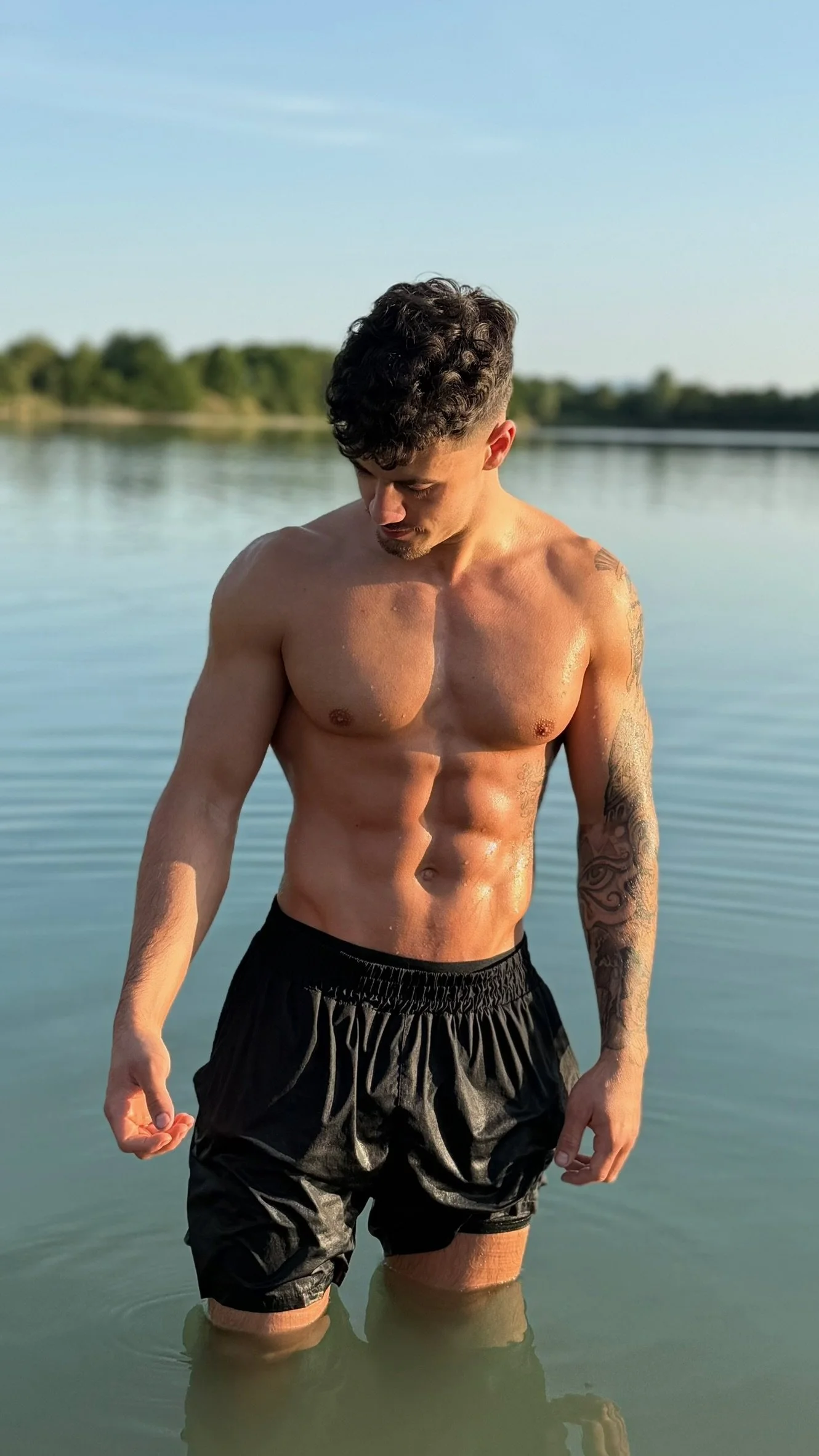 A muscular young man with tattoos on his arm, wearing black shorts, stands in a calm body of water during sunset with greenery and trees in the background.