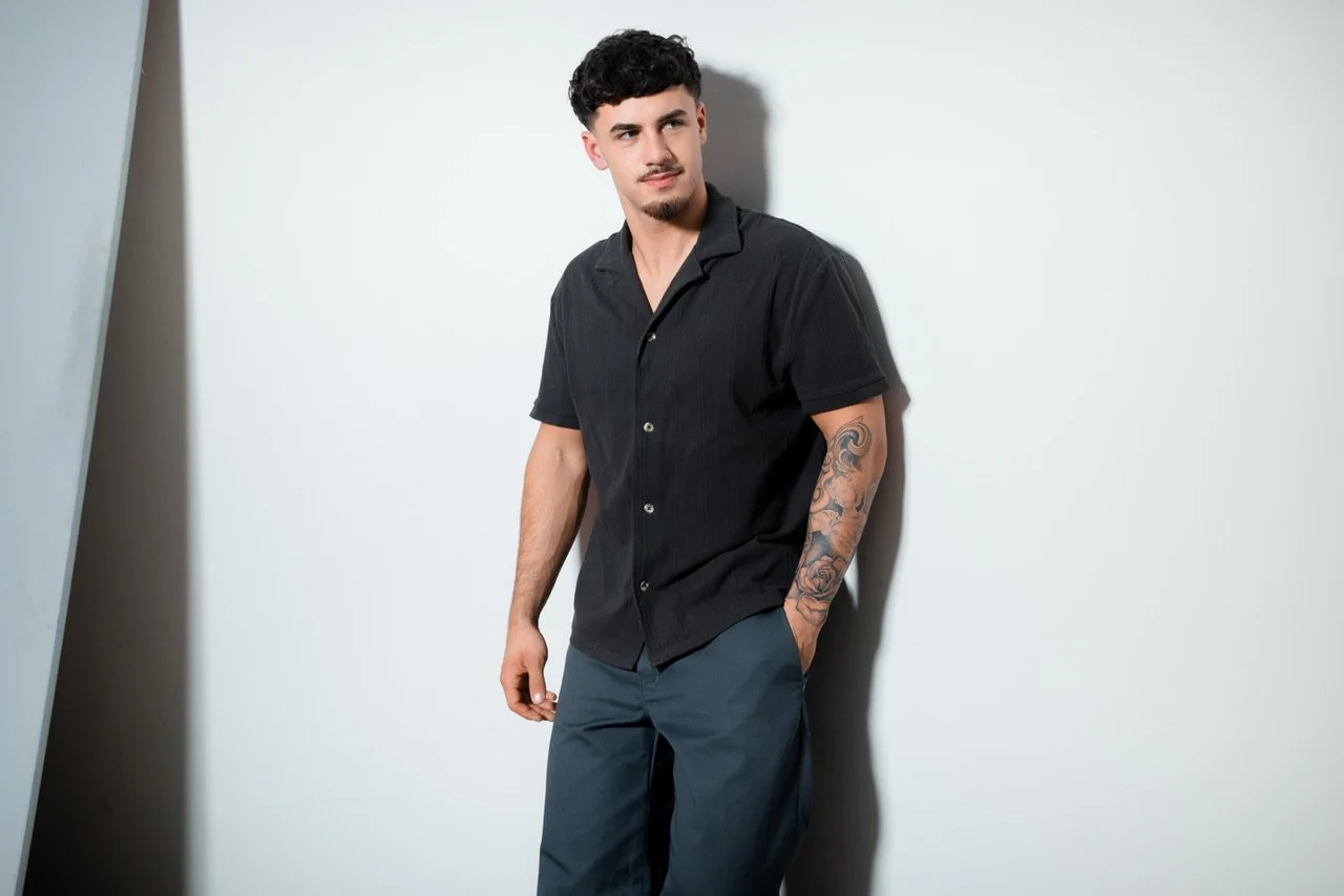 A young man with dark hair and a mustache, wearing a black short-sleeve button-up shirt and gray pants, standing against a plain white wall with one hand in his pants pocket and the other hanging down, looking to the side.