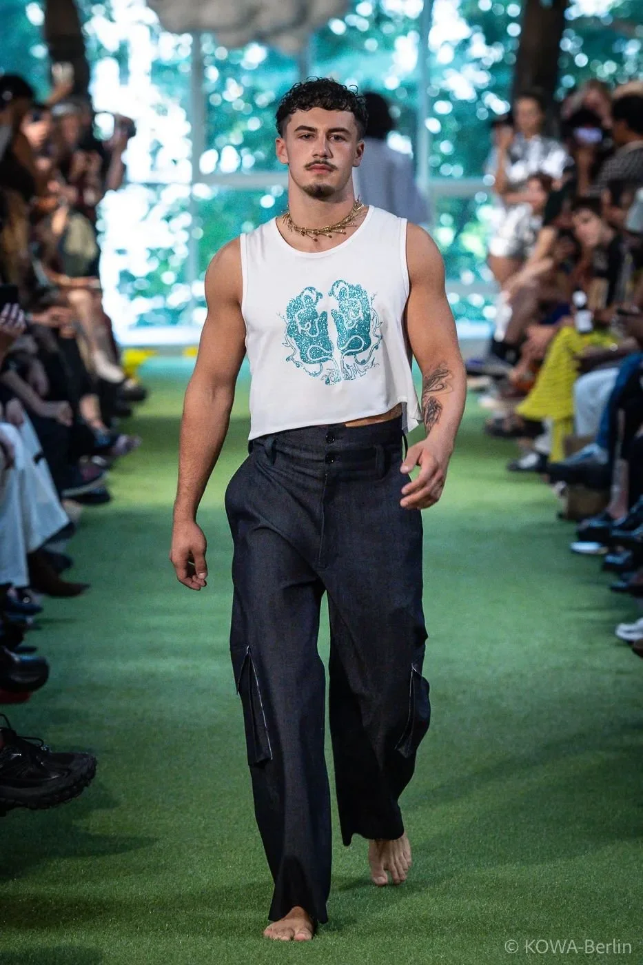 A young male model walking barefoot on a green runway during a fashion show, wearing a sleeveless white cropped tank top with blue graphics and loose black cargo pants, with an audience seated on both sides.