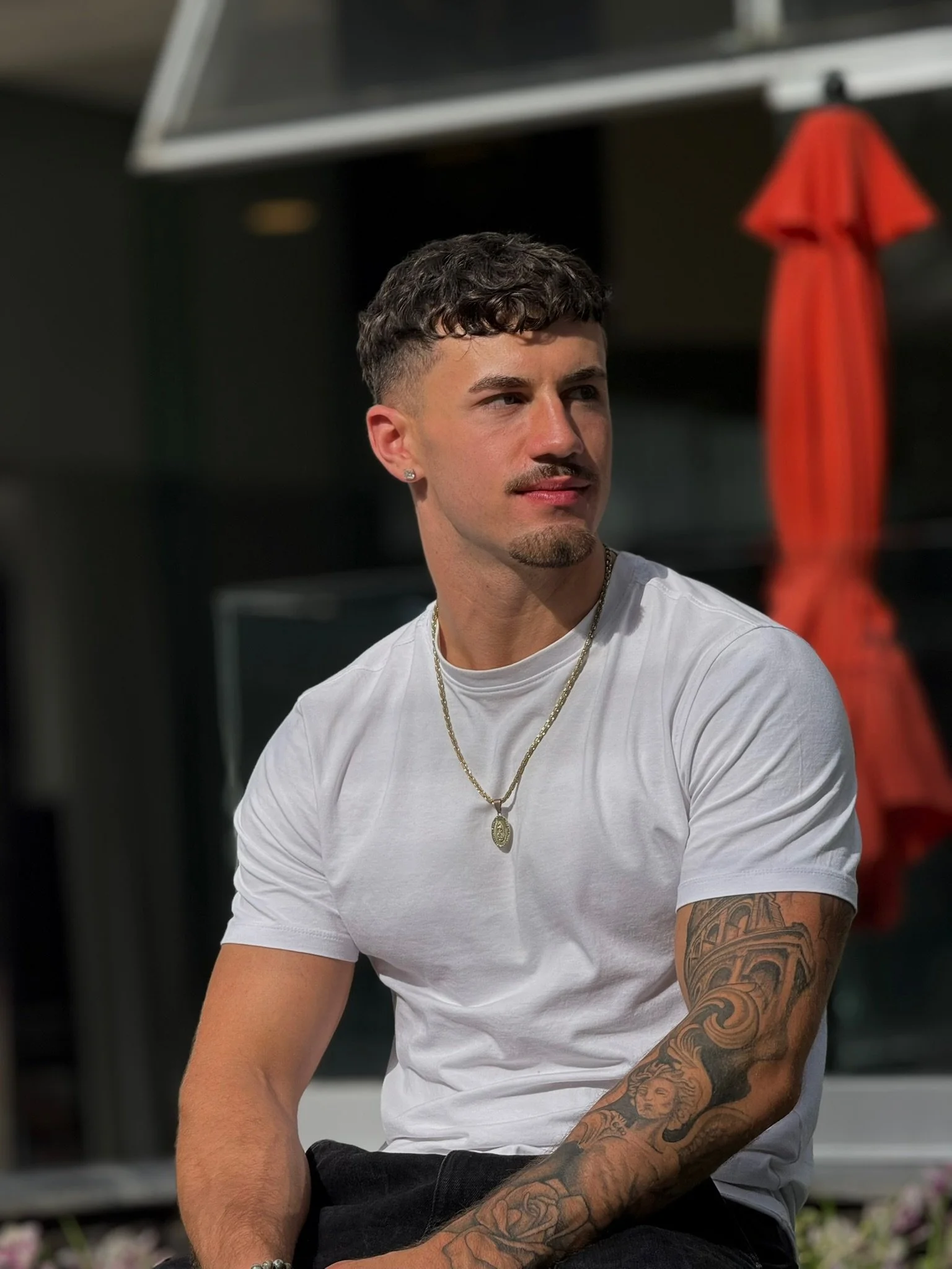 A young man with short curly hair, a mustache and goatee, wearing a white t-shirt, gold chain necklace, and silver earrings, sitting outdoors with a red umbrella in the background.