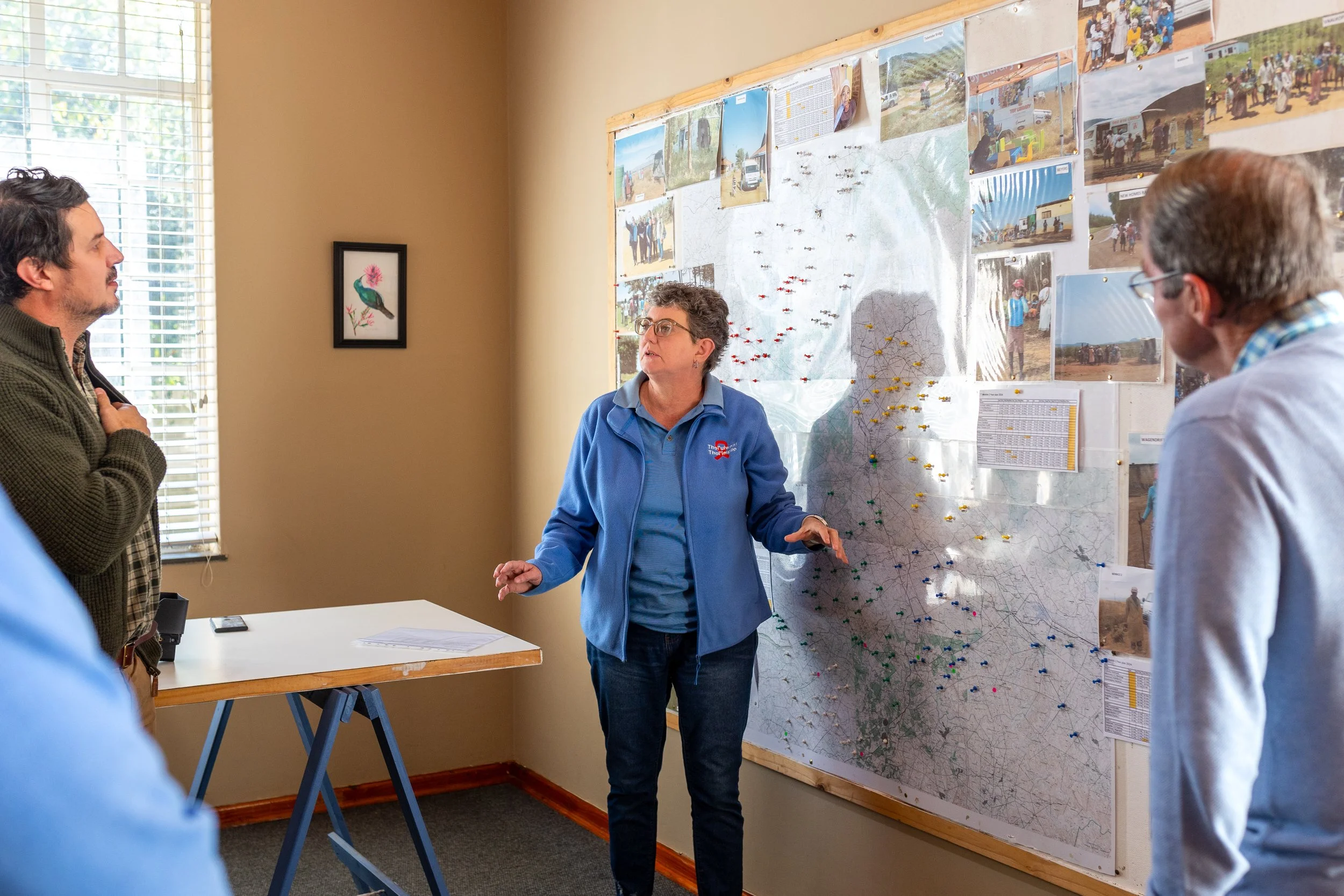 A woman s standing in front of a large map on a wall, explaining the TTM reach and mobile clinic routes to a group of people. The map has photos and colored pins. Three men are listening.