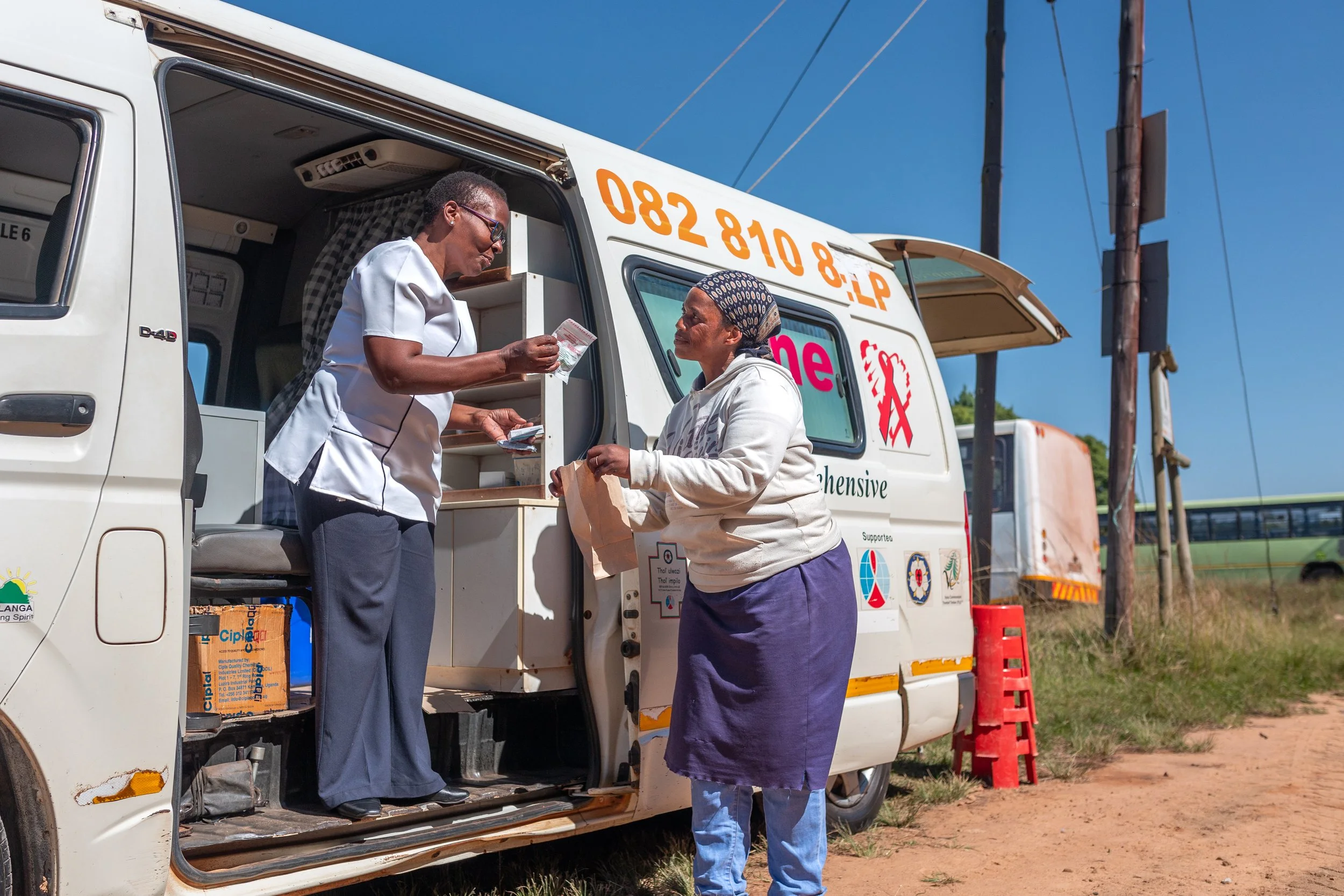 A mobile pharmacy van with two women, one inside handing medication to the other outside, on a dirt road with a blue sky and power lines in the background.