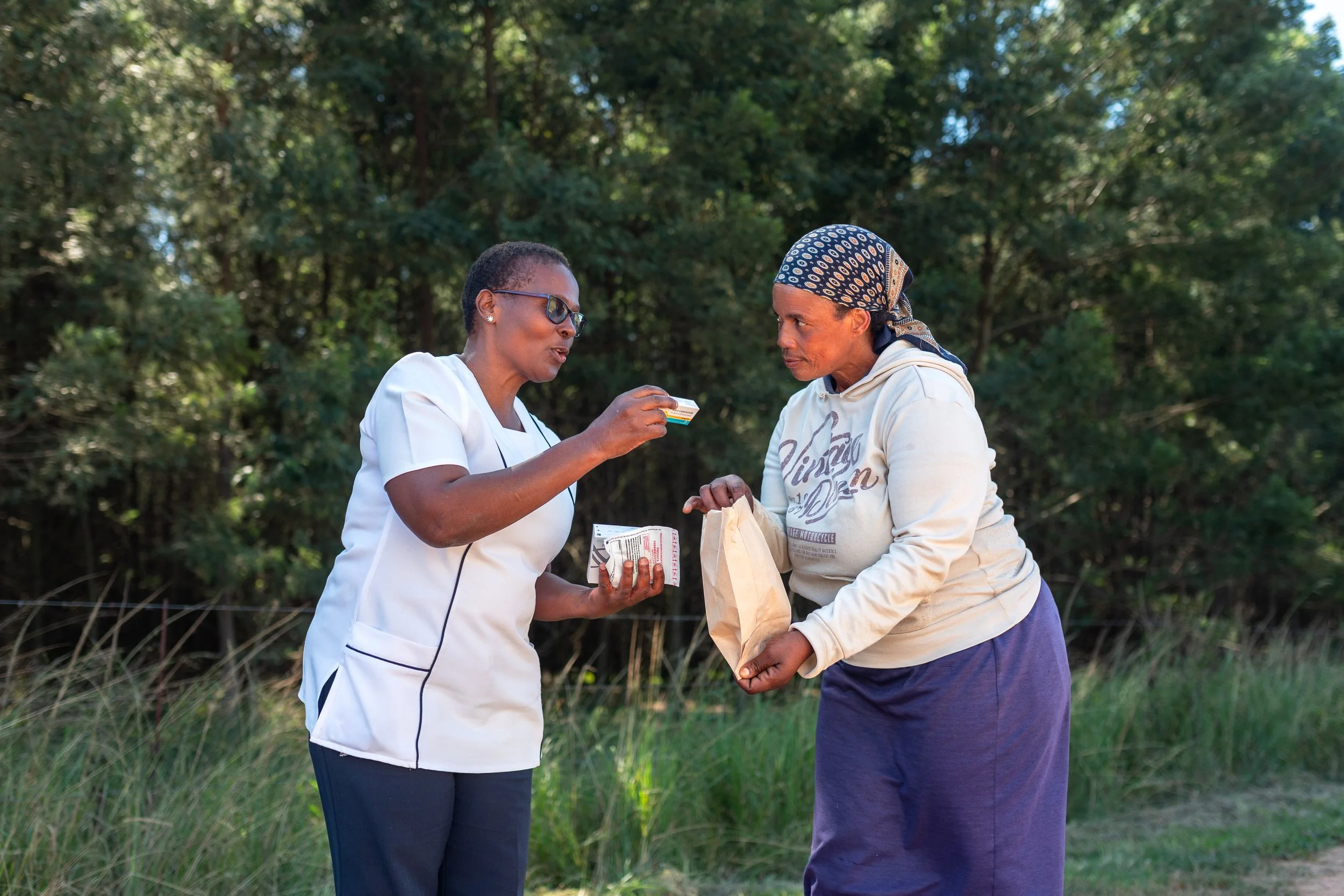 A healthcare worker in white scrubs hands over medication to another woman wearing a headscarf and hoodie outdoors in front of trees.