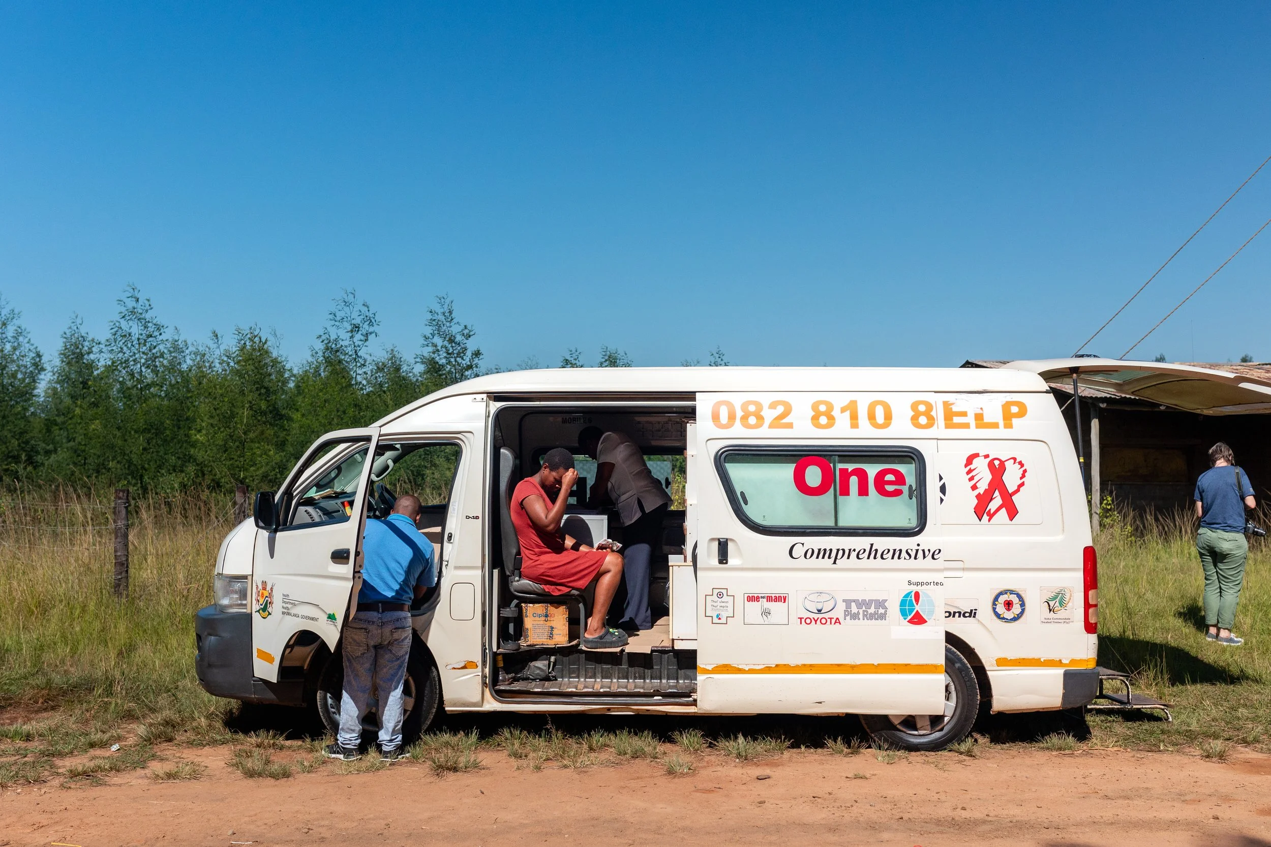 A white mobile clinic vehicle parked on a dirt road with three people around it, one seated inside and two outside. The vehicle has various logos and text, including a red ribbon symbol and the words "One" and "Comprehensive." One person is in a blue shirt and jeans, another is in a red dress sitting inside, and a third person is walking away on the right side.
