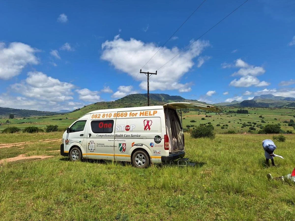 A white health care service van parked on a grassy field with hills and a blue sky with clouds in the background. The van has contact information and health-related logos on it, and a person is standing nearby with a bag.