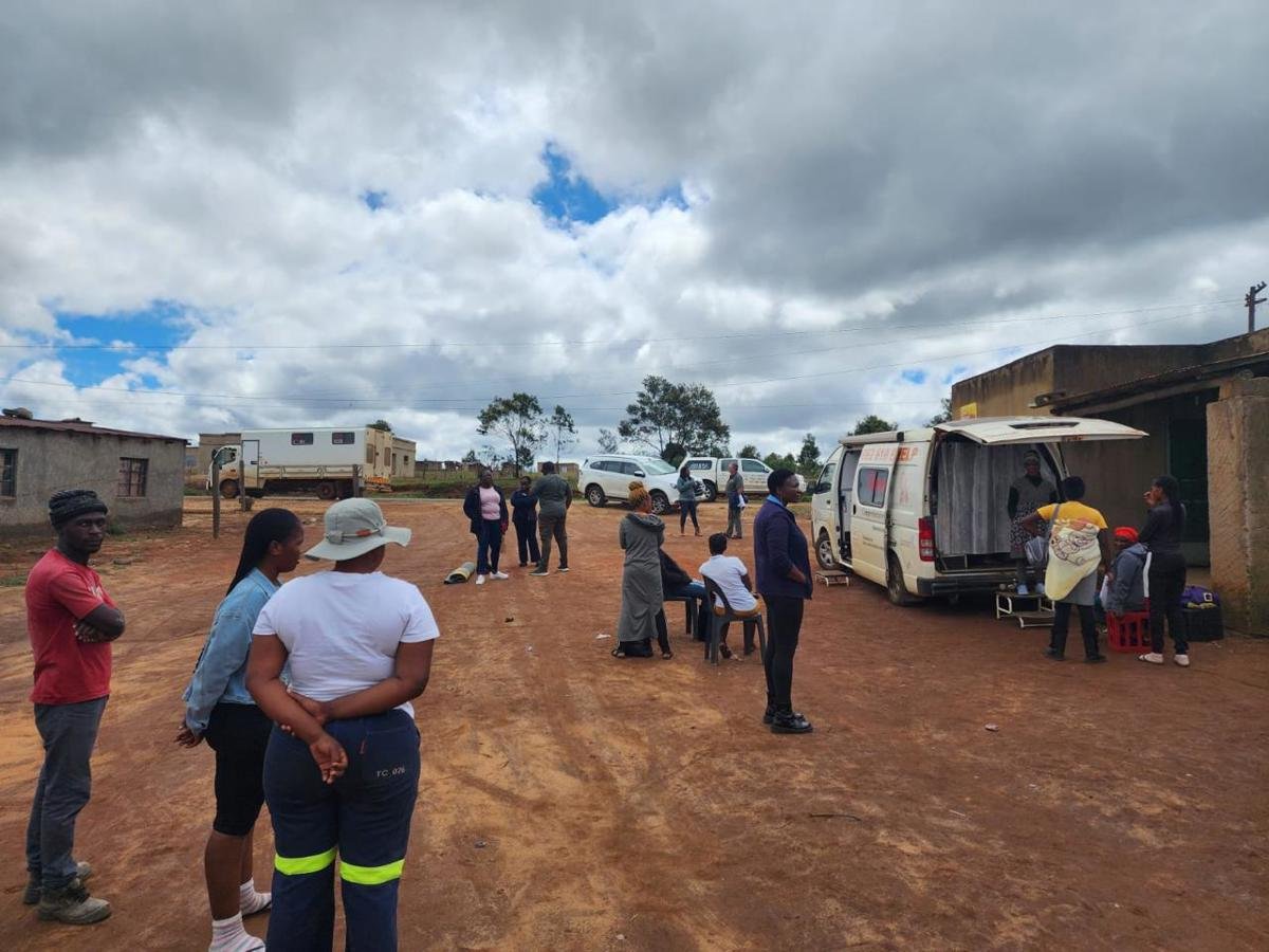People gather outside on a dirt area near a small building, with several vehicles including a van and a white SUV parked nearby. Some individuals are standing, sitting, or interacting near the vehicles, under a cloudy sky.