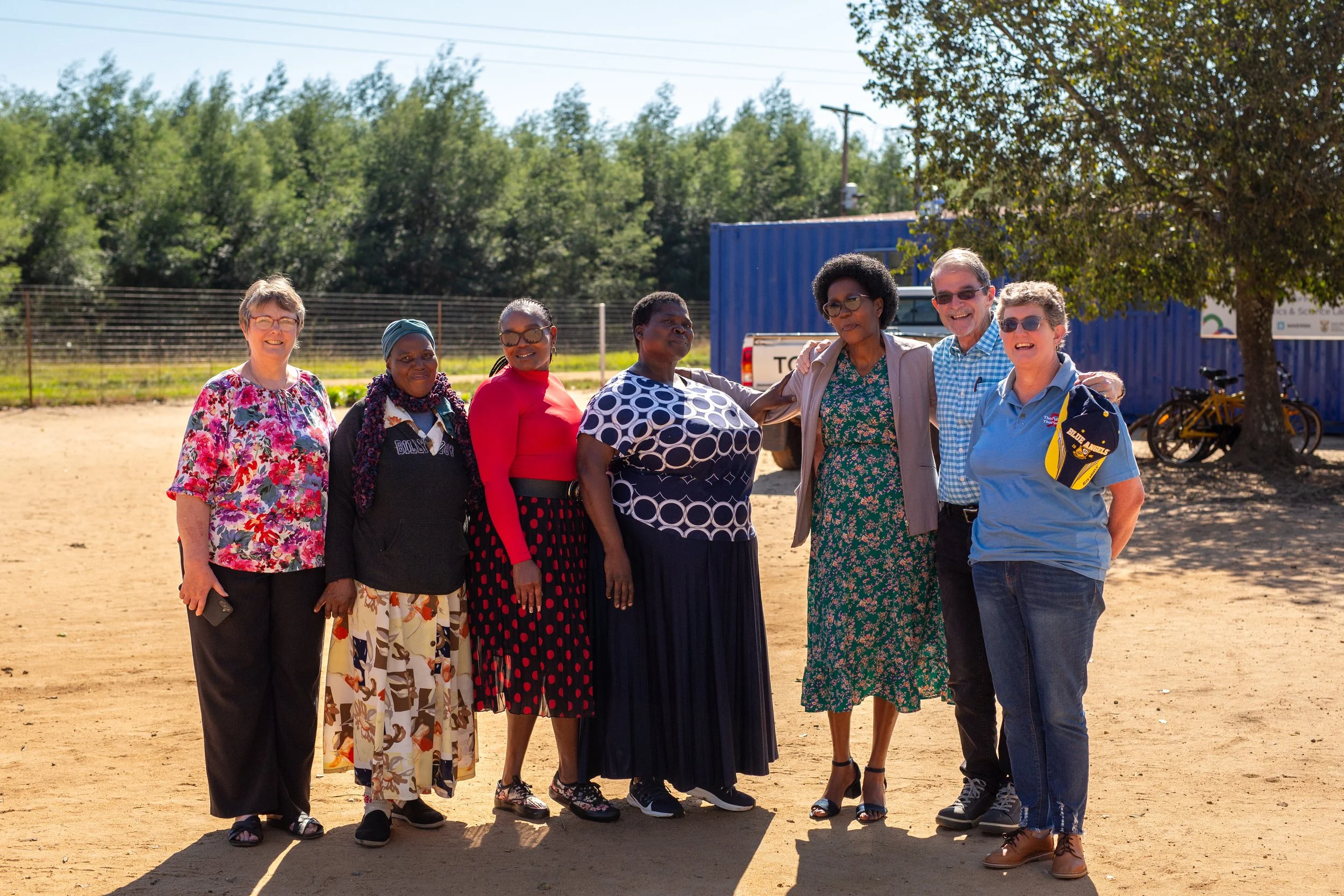 A group of eight diverse women and men standing outdoors on a sunny day, smiling and posing for the photo, with bicycles, a tree, and a blue truck in the background.