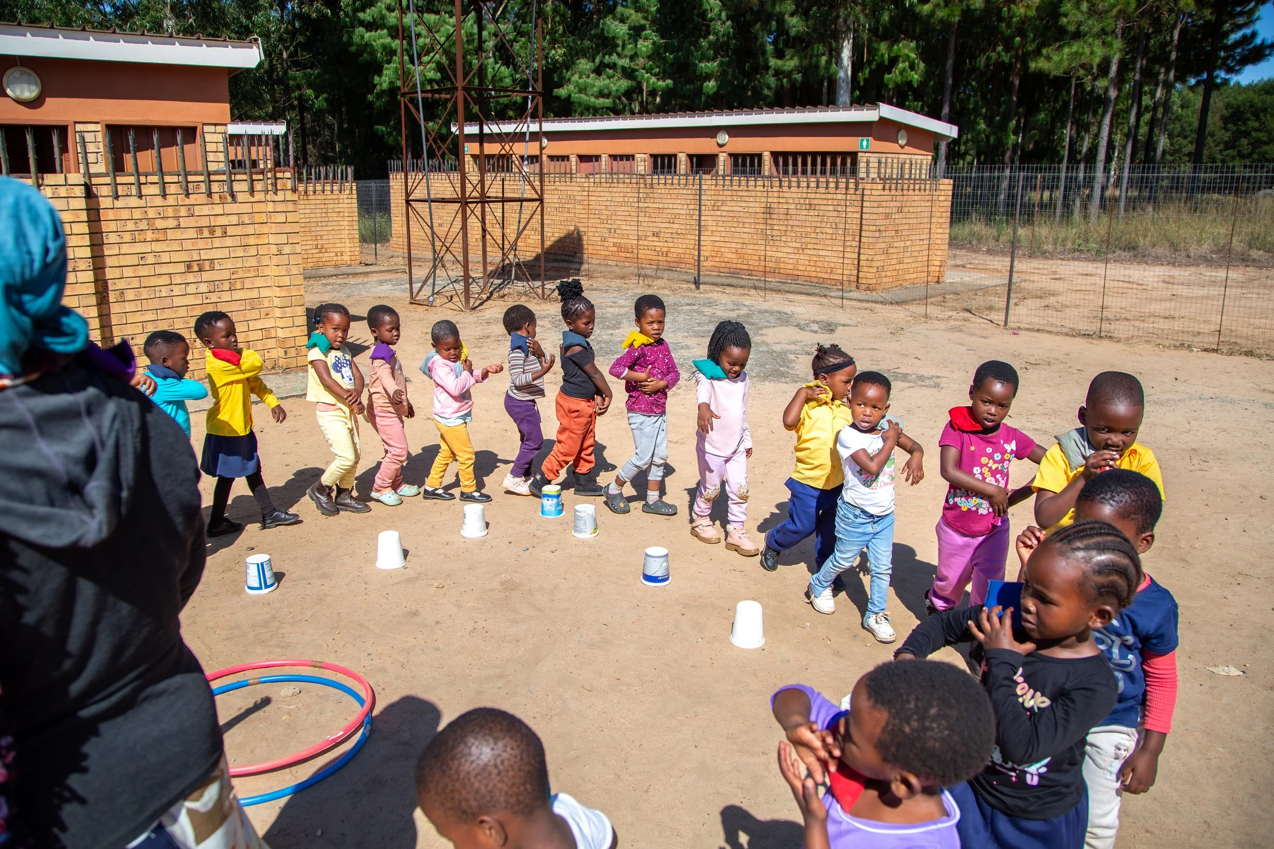 Children participating in an outdoor activity or game, forming a line and stepping over cups, supervised by an adult, on a sandy playground with a brick building and trees in the background.