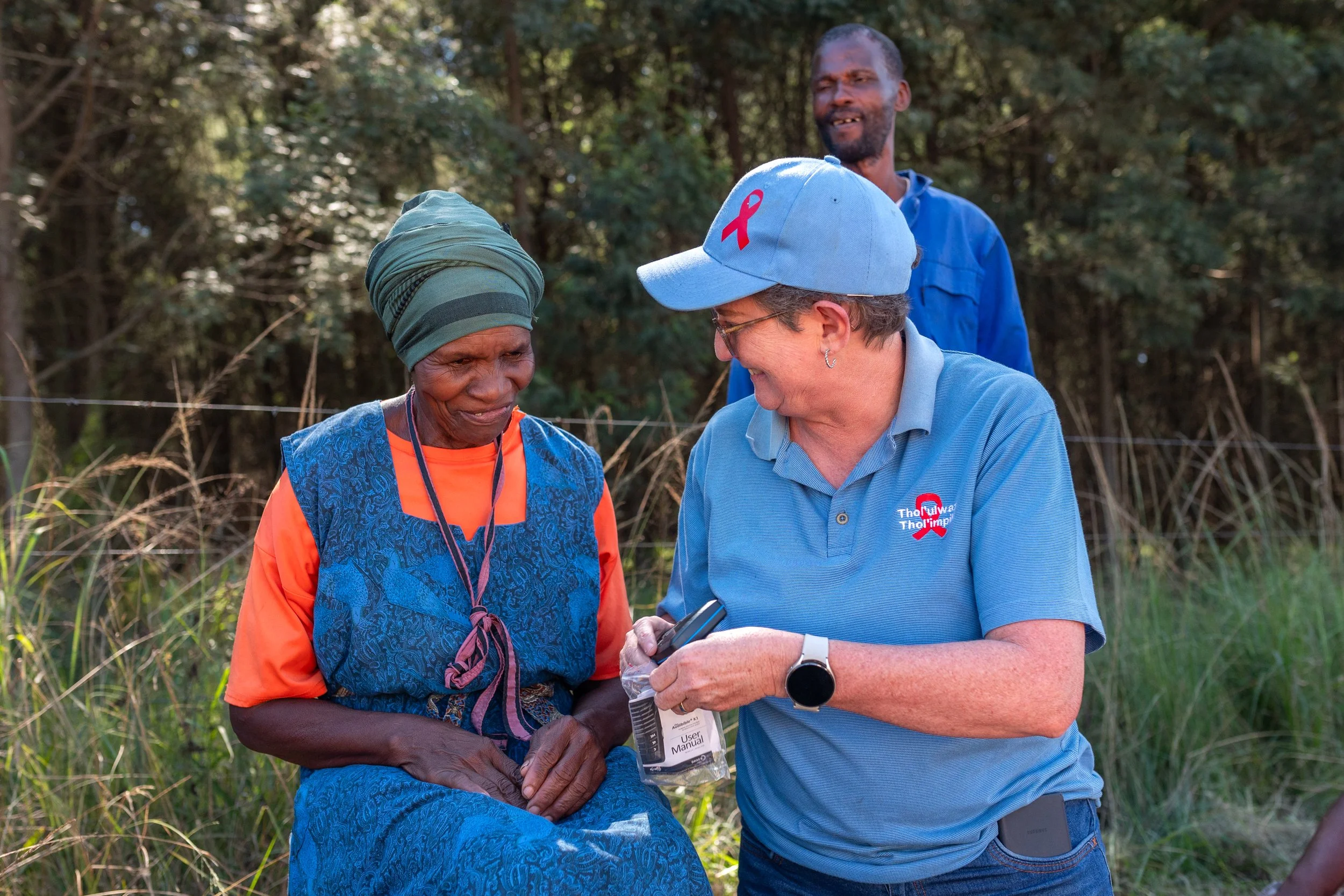 Two women and a man outdoors in a grassy, wooded area. The women are smiling and engaging. 
