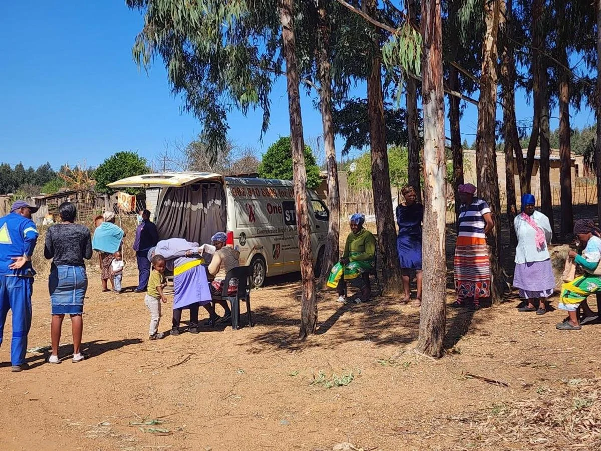 People gathering outdoors around a vehicle, sitting and standing under trees, with a dirt ground and wooden structures in the background on a sunny day.