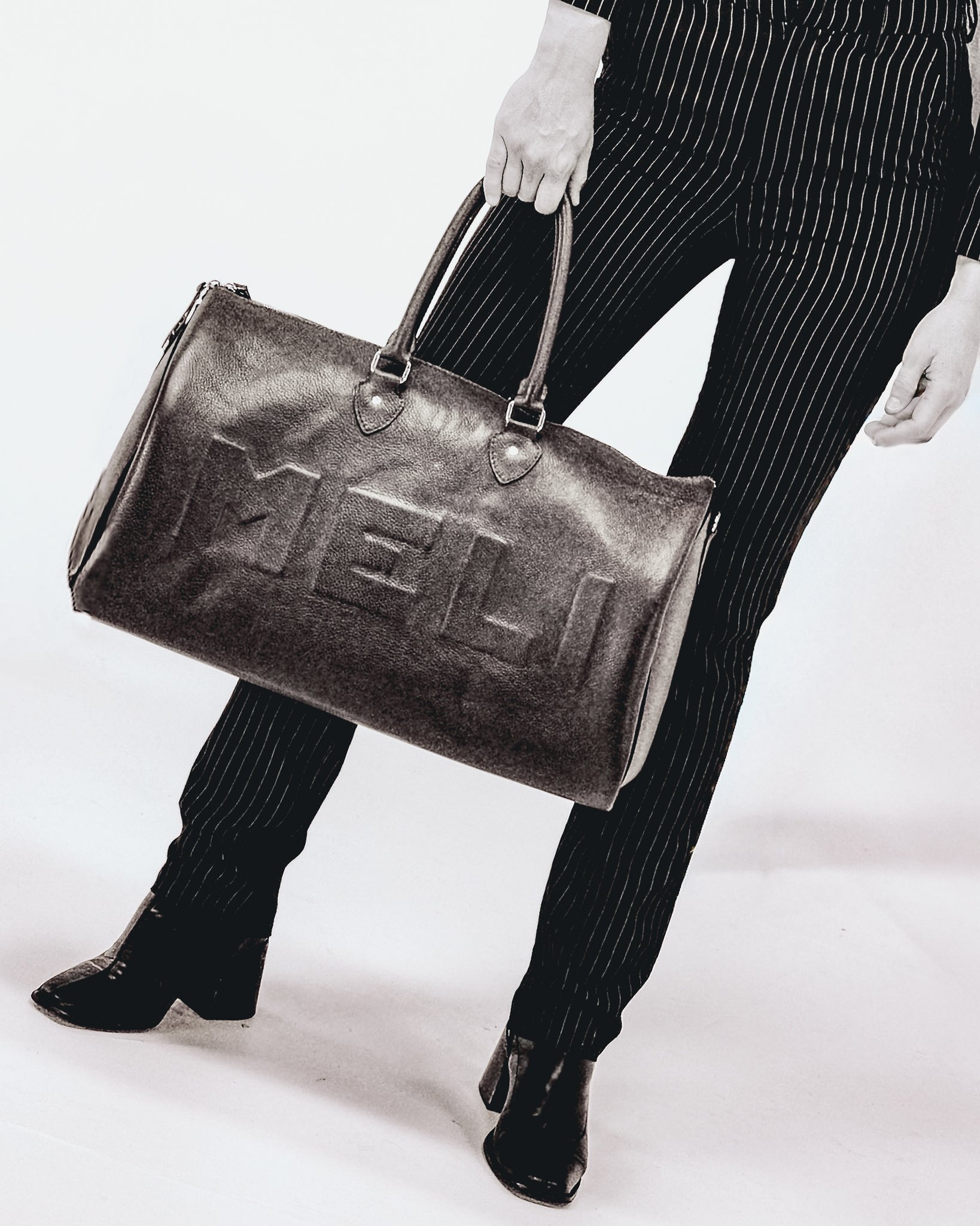 Person in black striped pants and black boots, holding a large leather handbag with the word 'FELLO' embossed on it, against a plain background.
