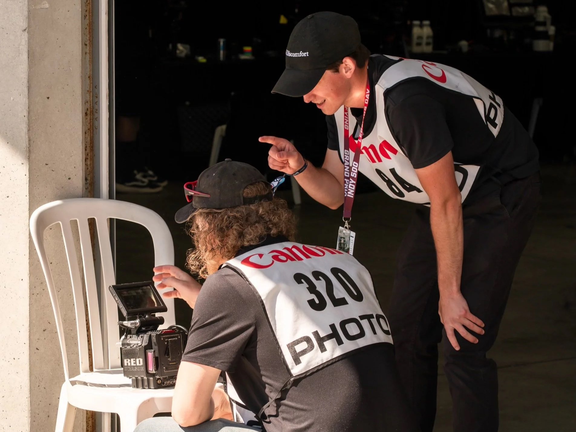 Two race officials or photographers, wearing black caps and black and gray uniforms with 'Canon' and 'PHOTO' written on vests, are working together near a white plastic chair. One is sitting and looking at a camera or recording device, while the othe