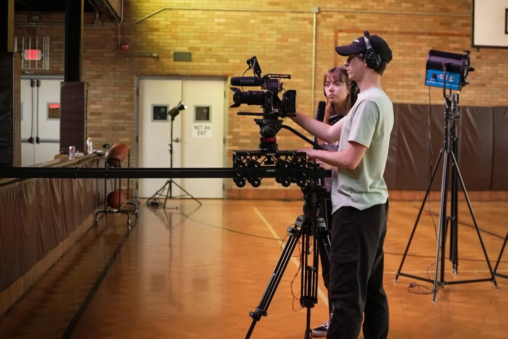Two camera operators filming in an indoor gymnasium with wooden floors and brick walls, positioned near the basketball court.