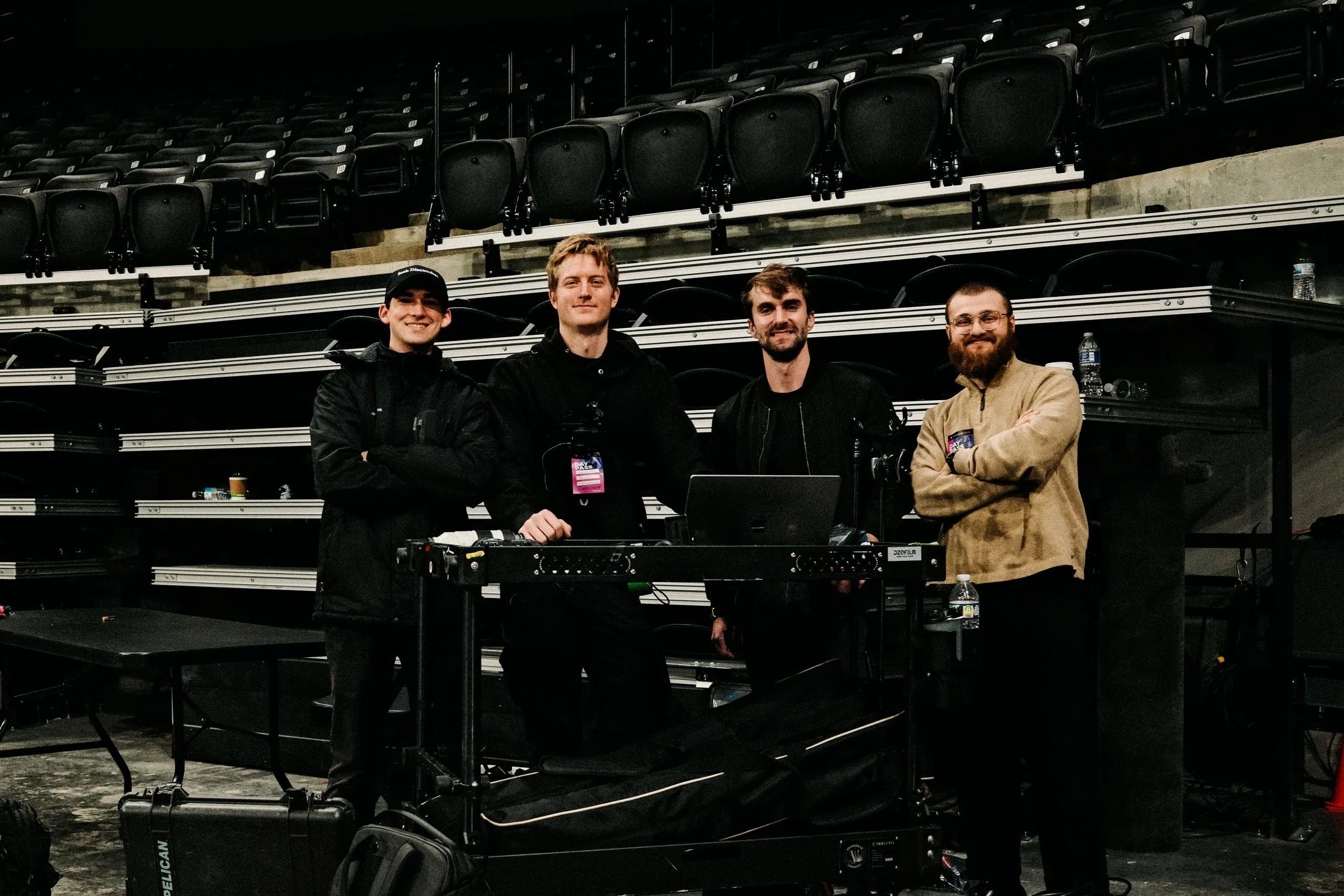Four men standing behind a table with equipment, in an indoor arena with empty black seats in the background.