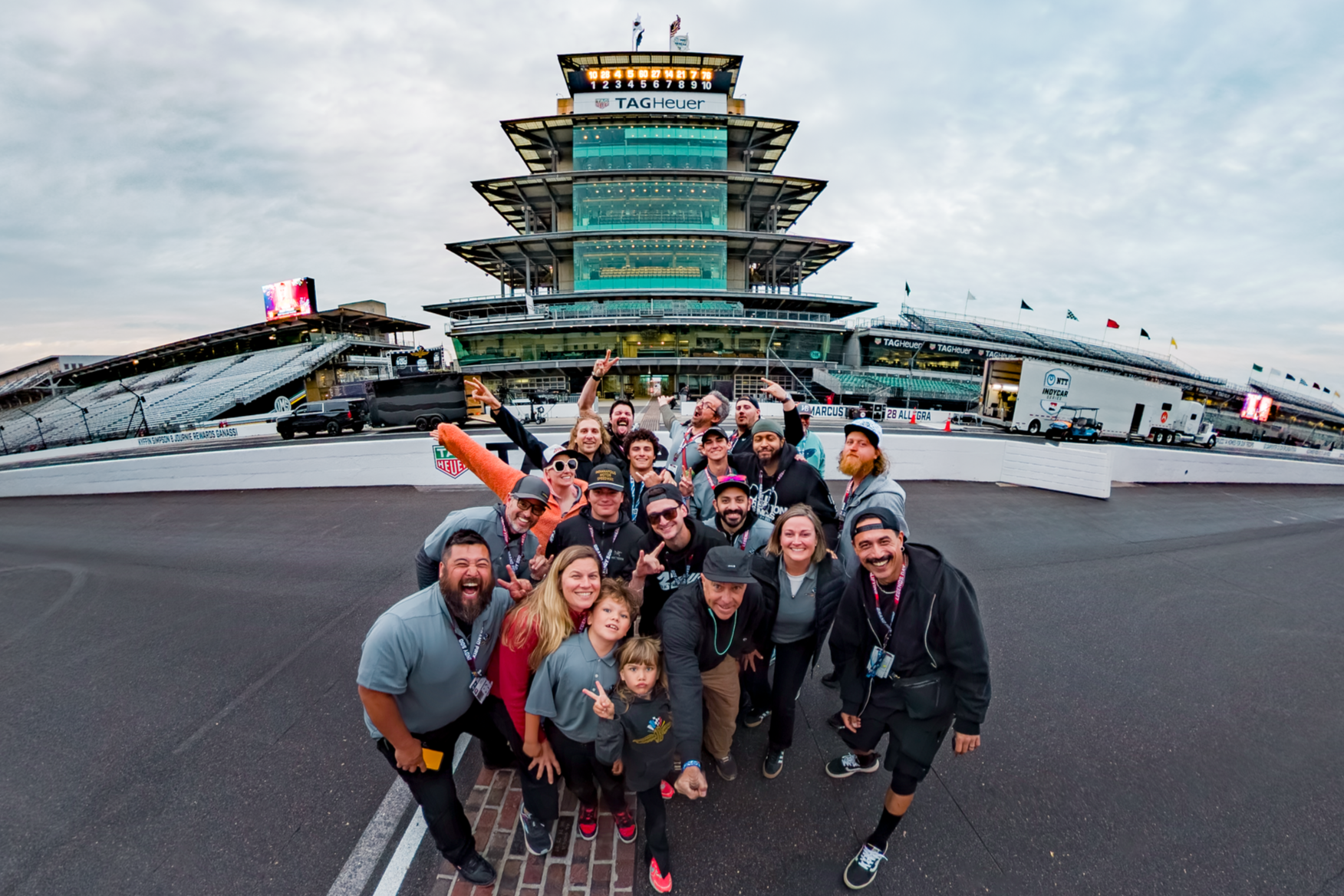 Group of people smiling and posing for a photo in front of a race track and a multi-story building, likely at a racing event.