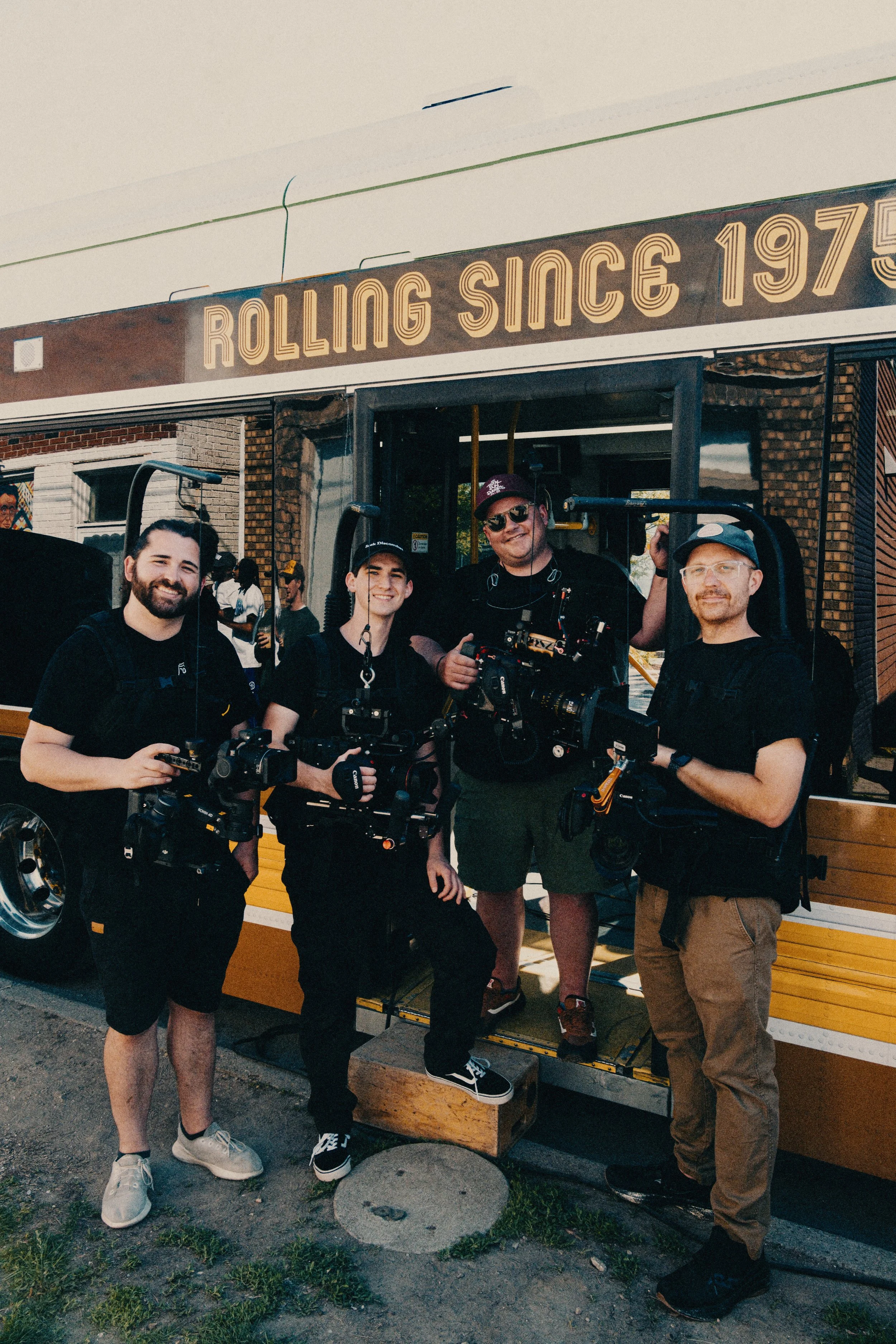 Four camera crew members standing in front of a vintage-style trolley with a sign that reads "Rolling Since 1975." They are holding professional filming equipment, smiling, and dressed casually.