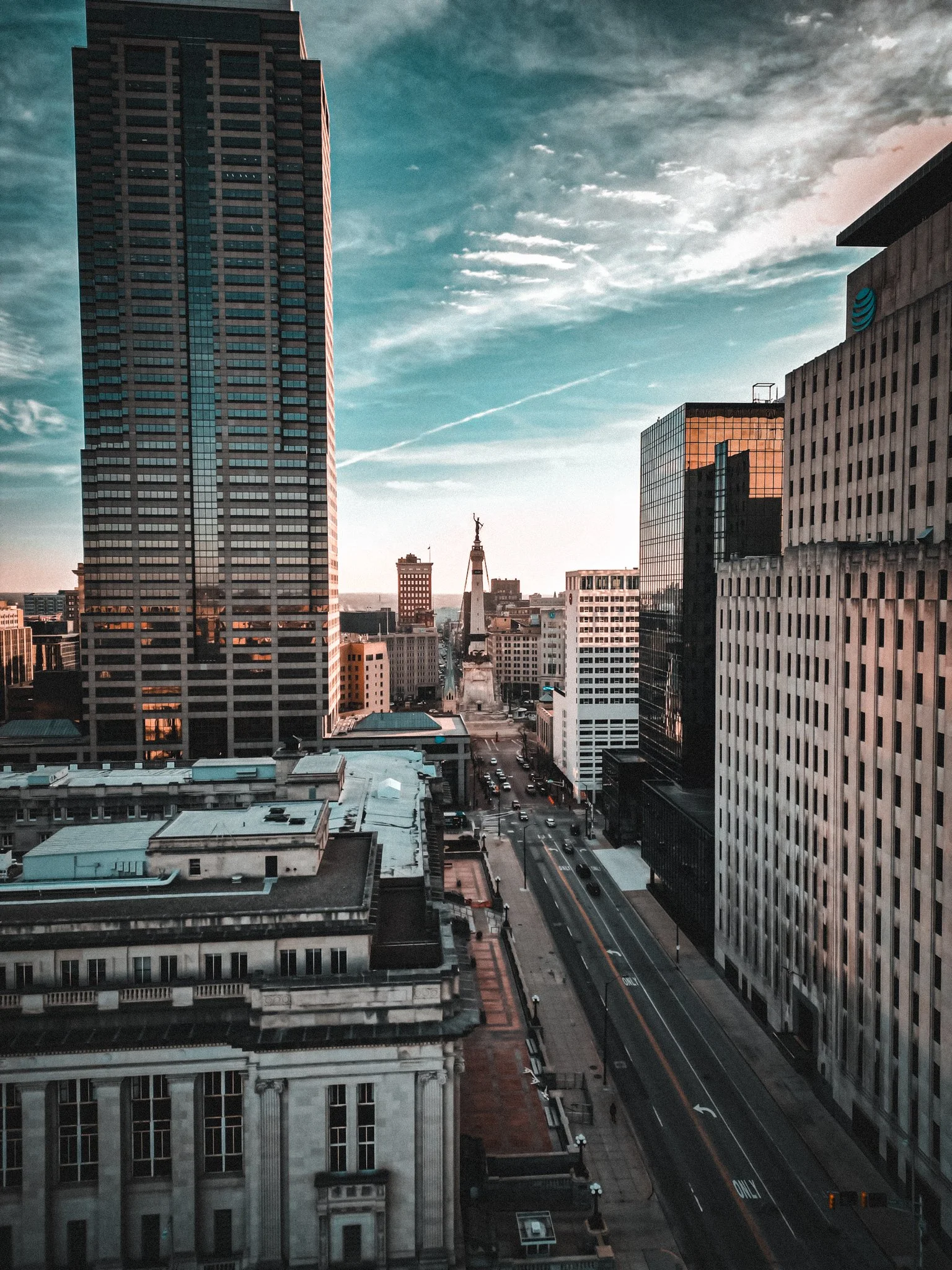 A cityscape at sunset featuring tall office buildings, a monument with a statue on top, and a street with few cars. The sky has clouds and contrails, with warm light reflecting off some buildings.