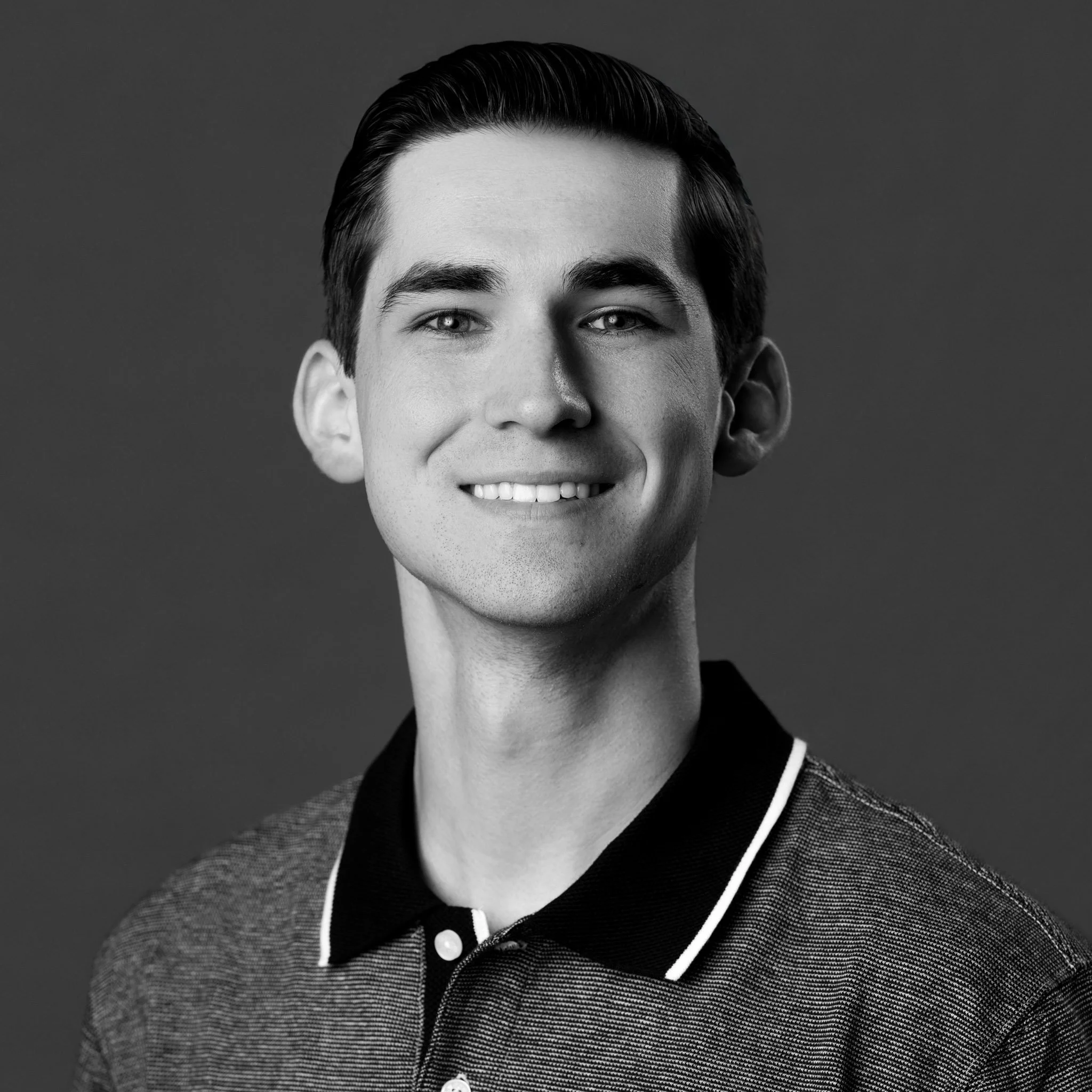 Black and white portrait of a young man with dark hair, smiling, wearing a collared shirt with a textured pattern.