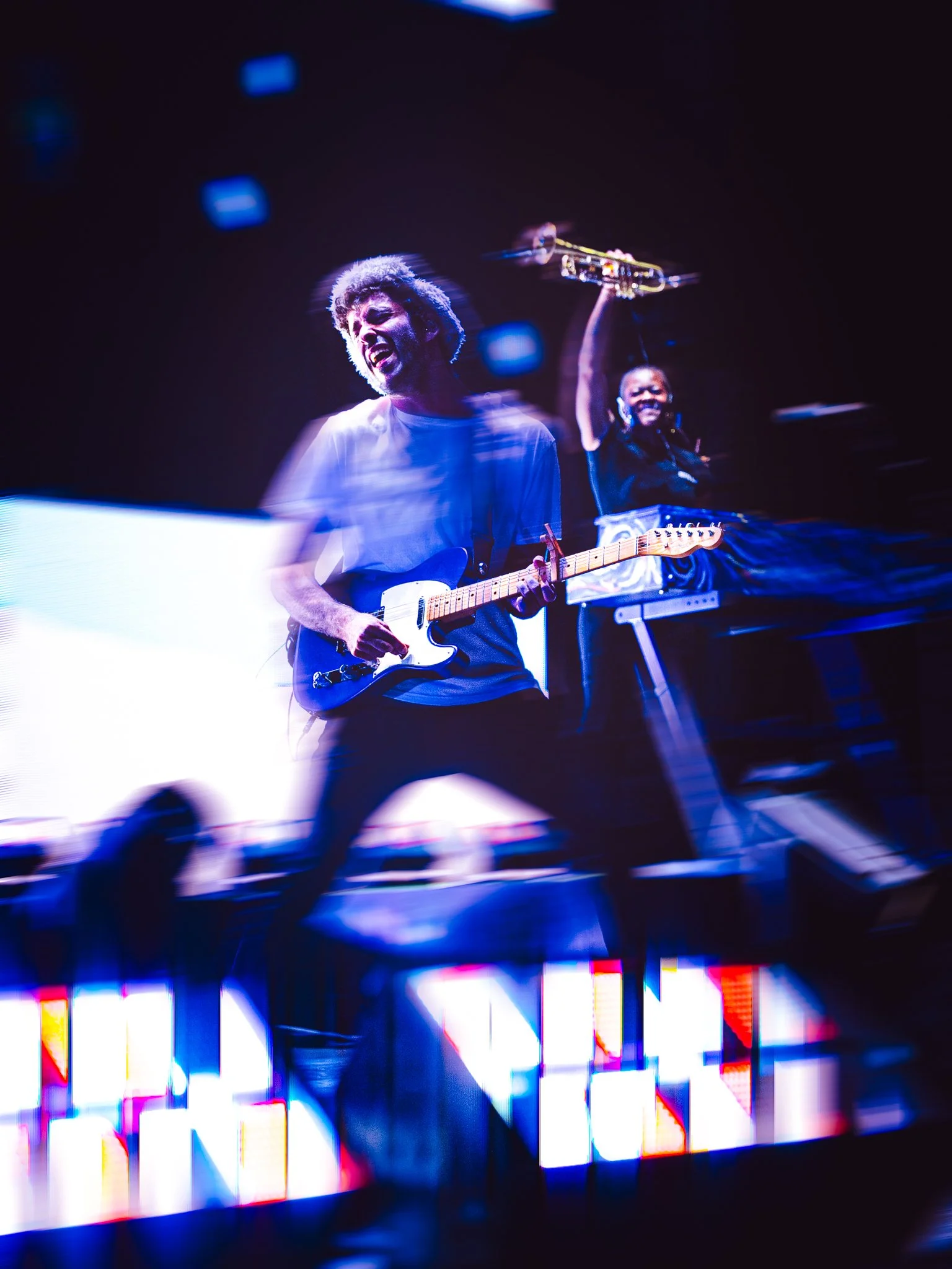 Two musicians performing on stage, one playing an electric guitar and singing passionately, the other holding a keyboard and raising a drumstick, with vibrant stage lights and digital screens in the background.