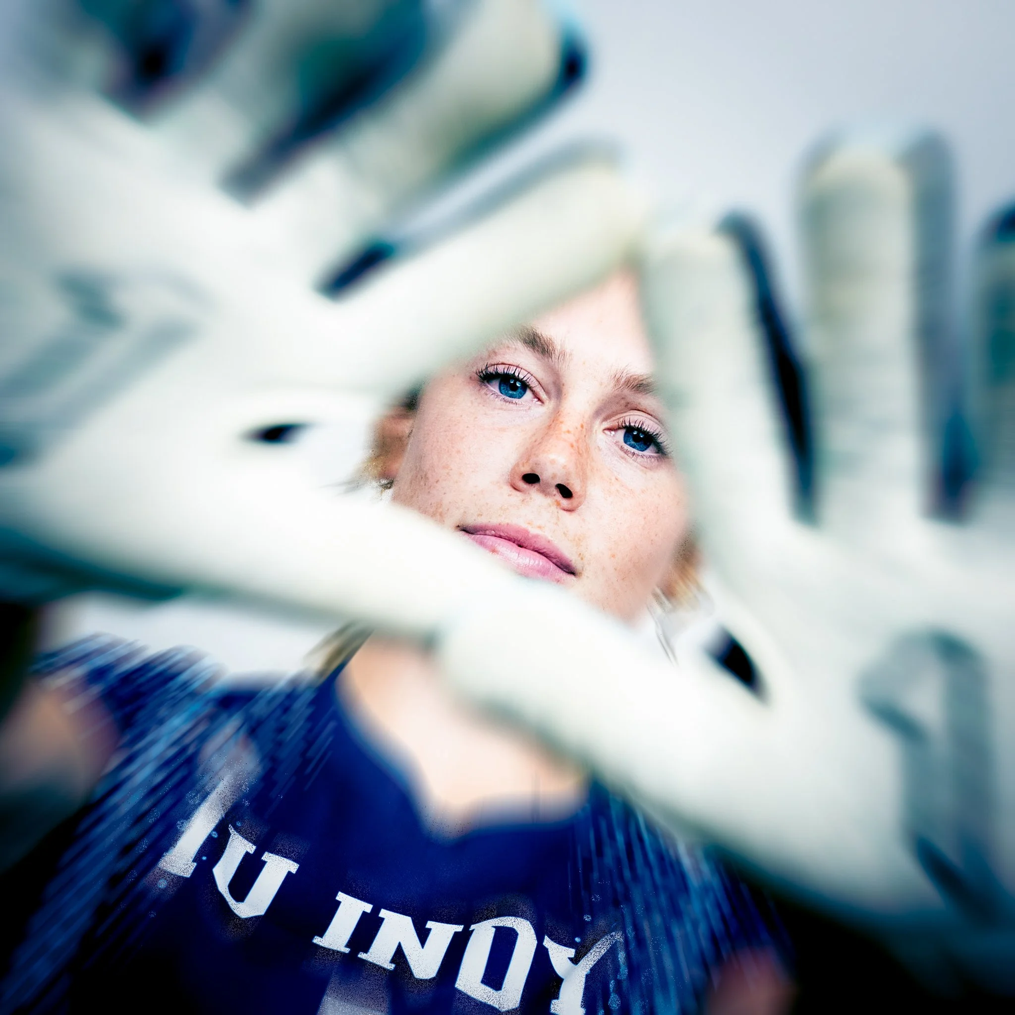 Close-up of a woman with blue eyes looking through a gap between gloved hands, wearing a dark shirt.
