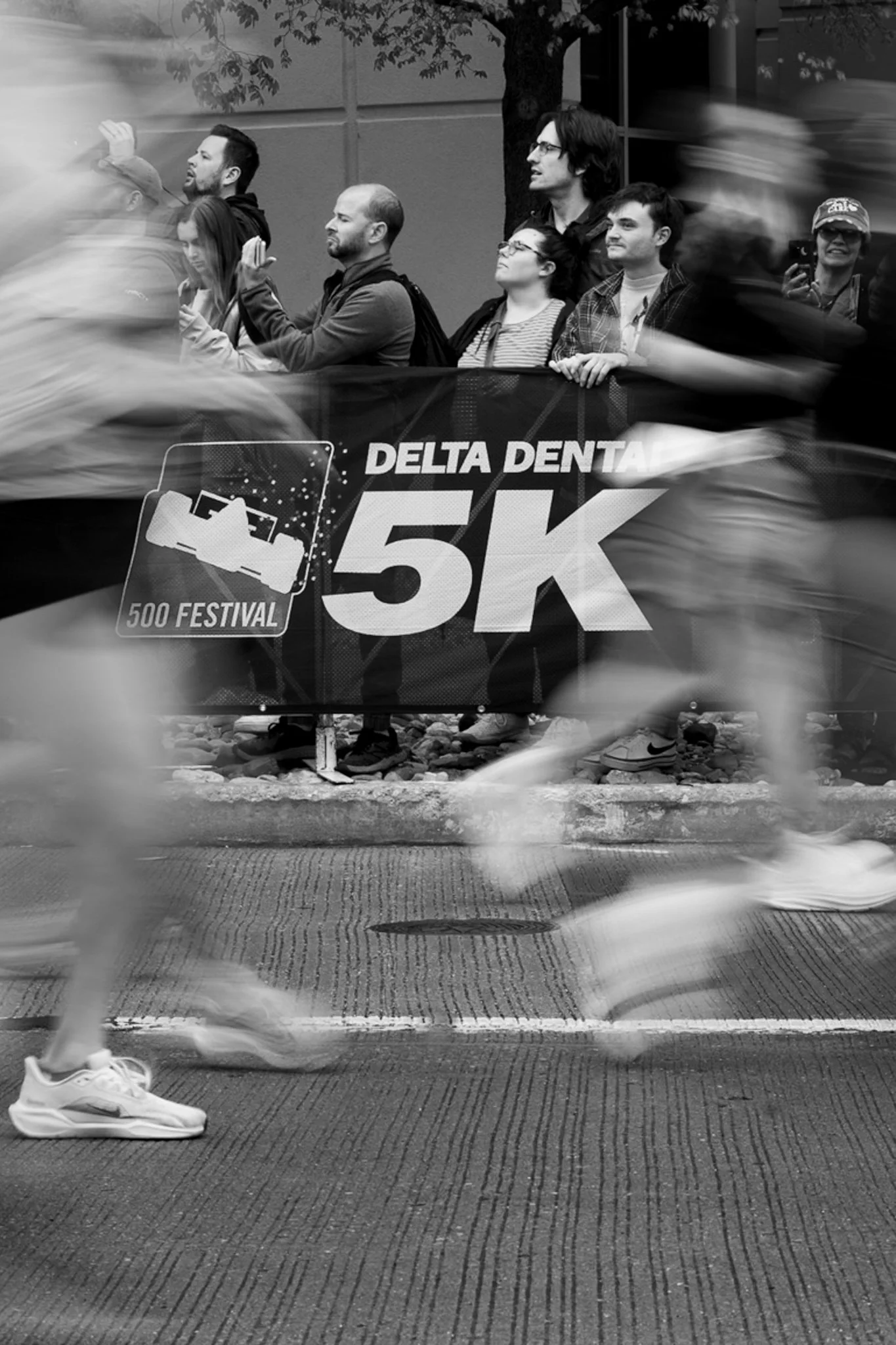 Participants running in the Delta Dental 5K race, with some spectators and a backdrop of trees and buildings.