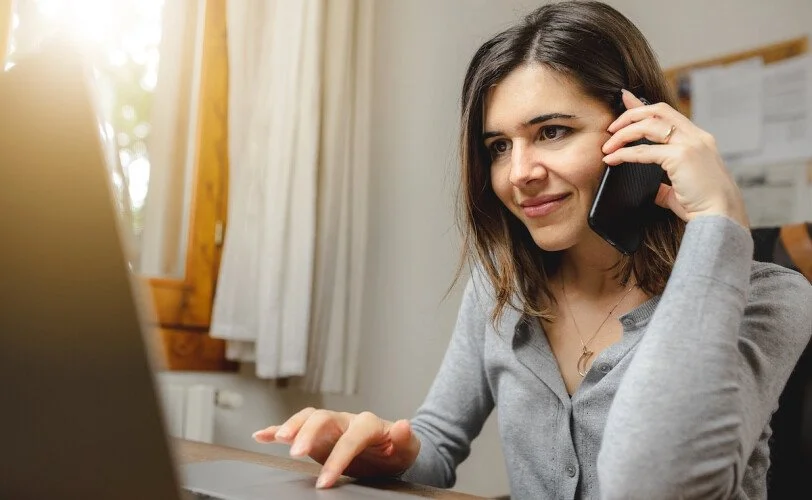 A woman with shoulder-length brown hair, wearing a gray shirt, is talking on a smartphone while working on a laptop in a well-lit room.