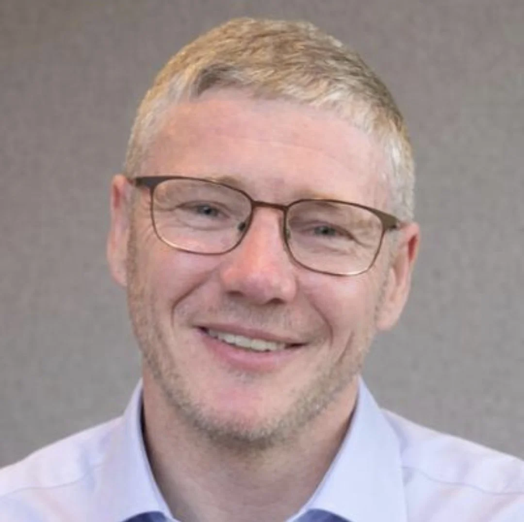 A smiling man with blonde hair, wearing glasses and a light blue shirt, against a neutral gray background.