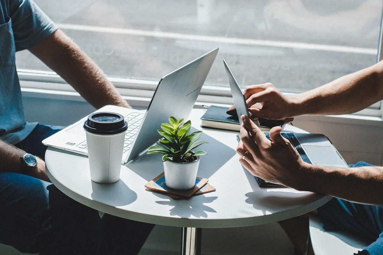Zwei Personen arbeiten an einem runden Tisch mit Laptop, Smartphone, Notizbuch, Kaffeebecher und einer Pflanze in einem hellen Raum neben einem Fenster.