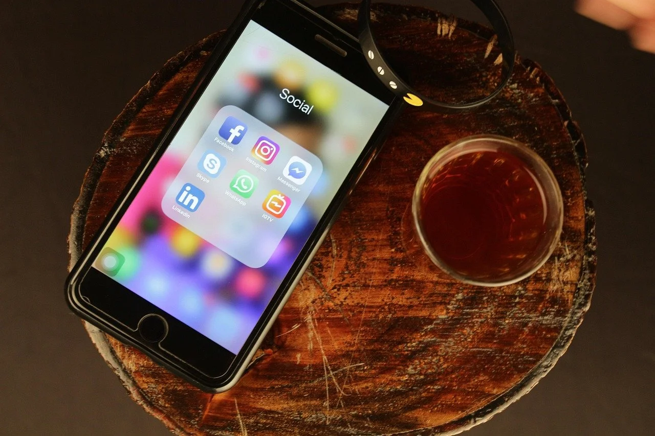 Smartphone mit Social-Media-Apps auf dem Bildschirm, daneben eine Tasse mit Getränk, auf einem runden Holztisch.
