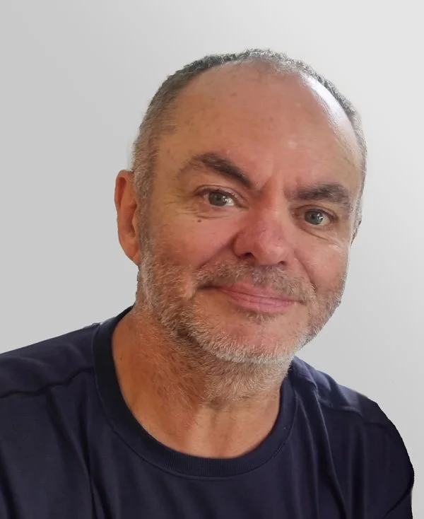 Close-up of a middle-aged man with gray hair and beard smiling, wearing a black shirt against a plain wall background.