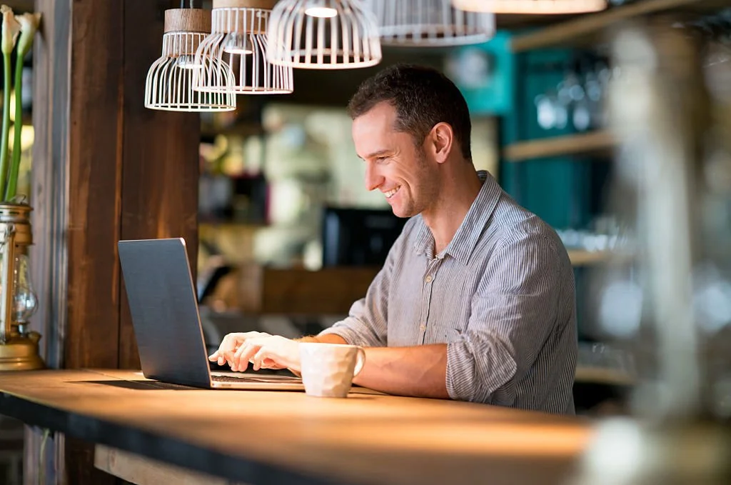A man sitting at a wooden counter in a cozy cafe, smiling while typing on a laptop, with hanging lamps above and a blurred background of shelves.