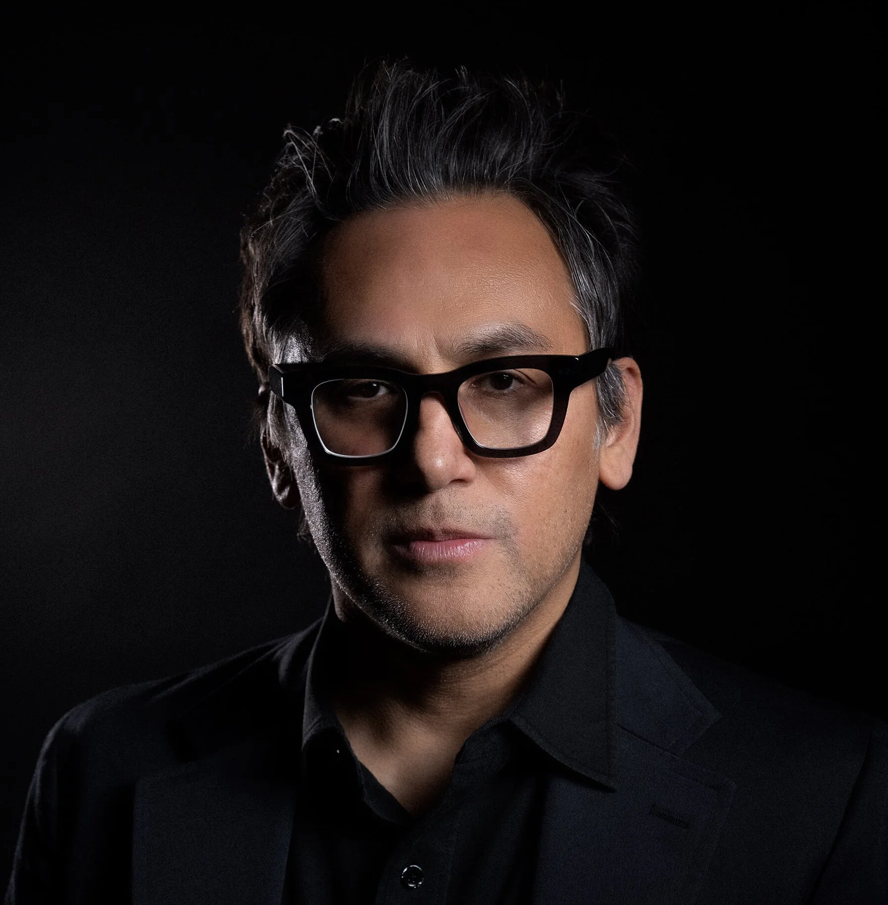 Portrait of Andrew Troy with black hair wearing black glasses and a black blazer, set against a dark background.