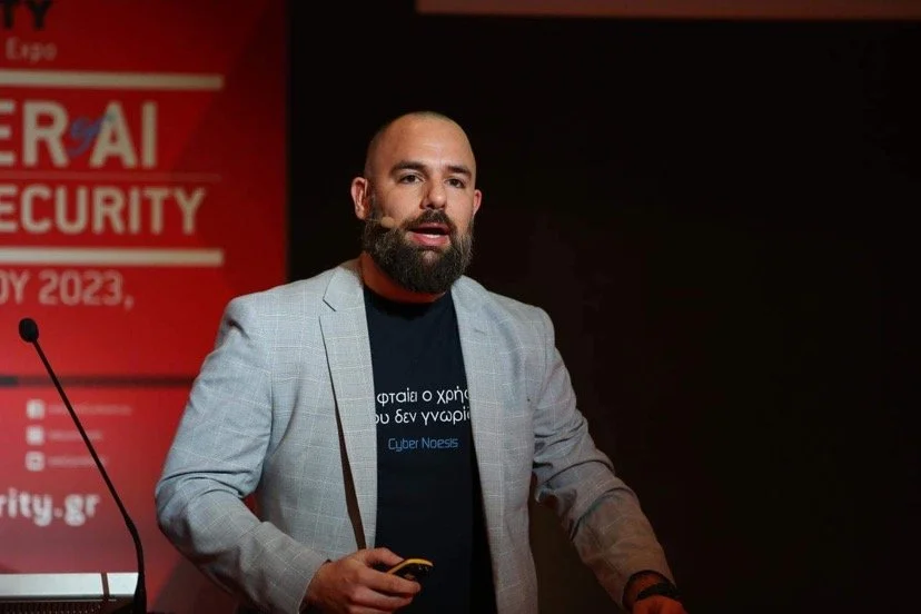 A man with a beard and bald head wearing a light gray blazer and dark t-shirt speaking at a conference stage with a red background signage related to AI security.