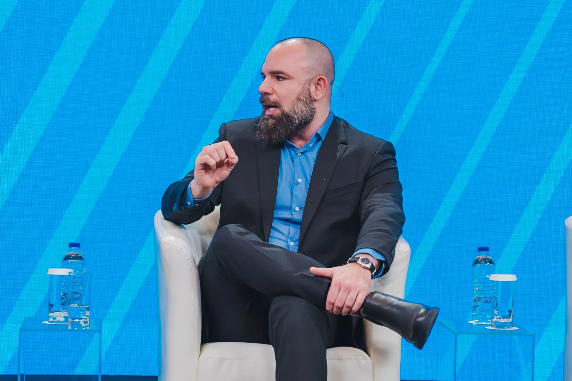 A bearded man with a shaved head in a dark suit and blue shirt, sitting in a white armchair on a stage, speaking into a microphone during a panel discussion. There are small tables on either side of him with water bottles and glasses. The background is a blue gradient with diagonal lines.