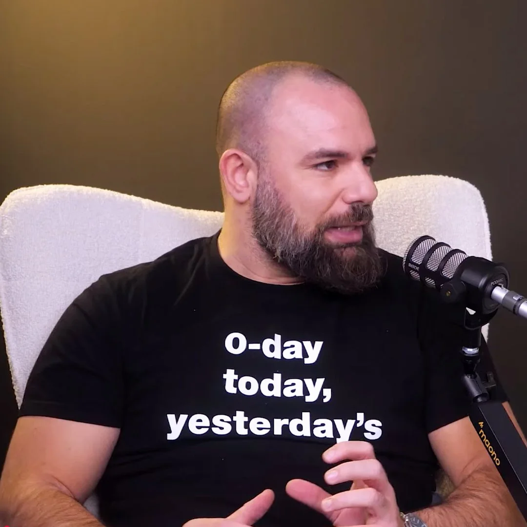 A man with a beard and shaved head sitting in a white chair speaking into a microphone, wearing a black T-shirt with the words 'o-day today, yesterday's' printed on it.