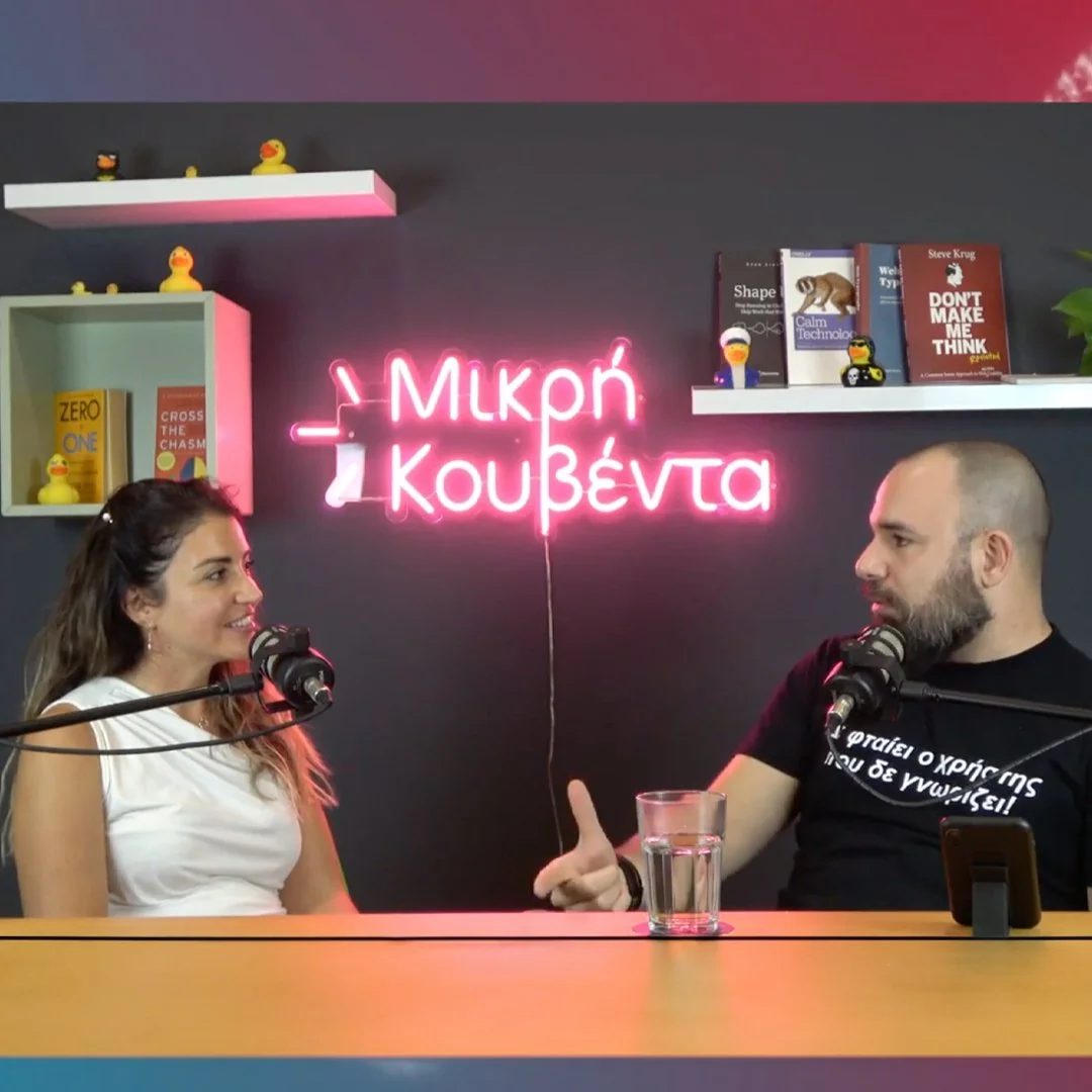 A woman and a man are sitting at a table with microphones in front of them, engaging in a conversation or podcast. The background features a dark wall with a pink neon sign in Greek that reads 'Μικρή κουβέντα,' and shelves with books, duck figurines, and small toys.
