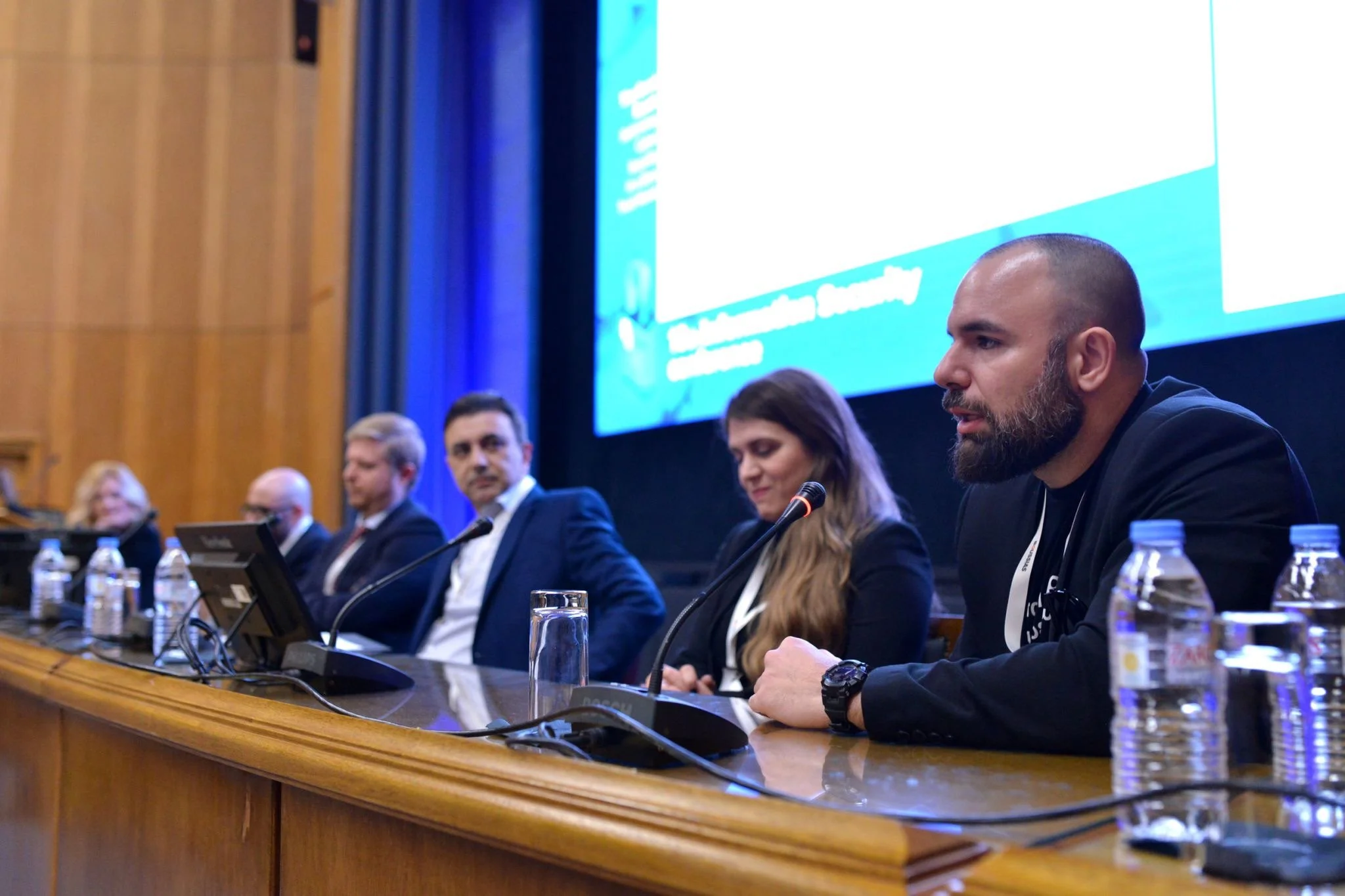 Panel of five people sitting at a long conference table, with microphones and water bottles, in front of a large screen displaying a presentation.