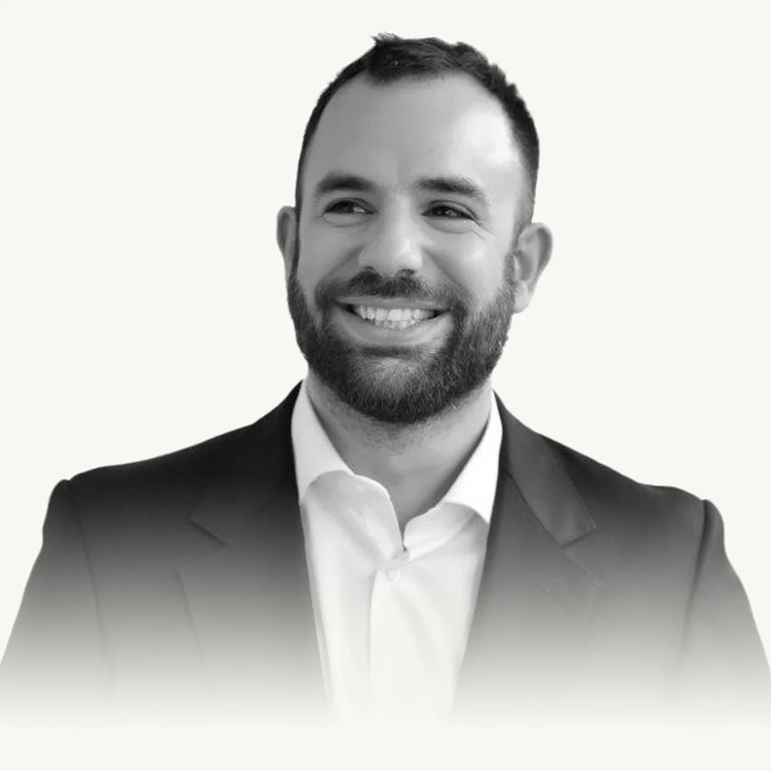 Black and white portrait of a smiling man with a beard, wearing a suit and white shirt, against a plain background.