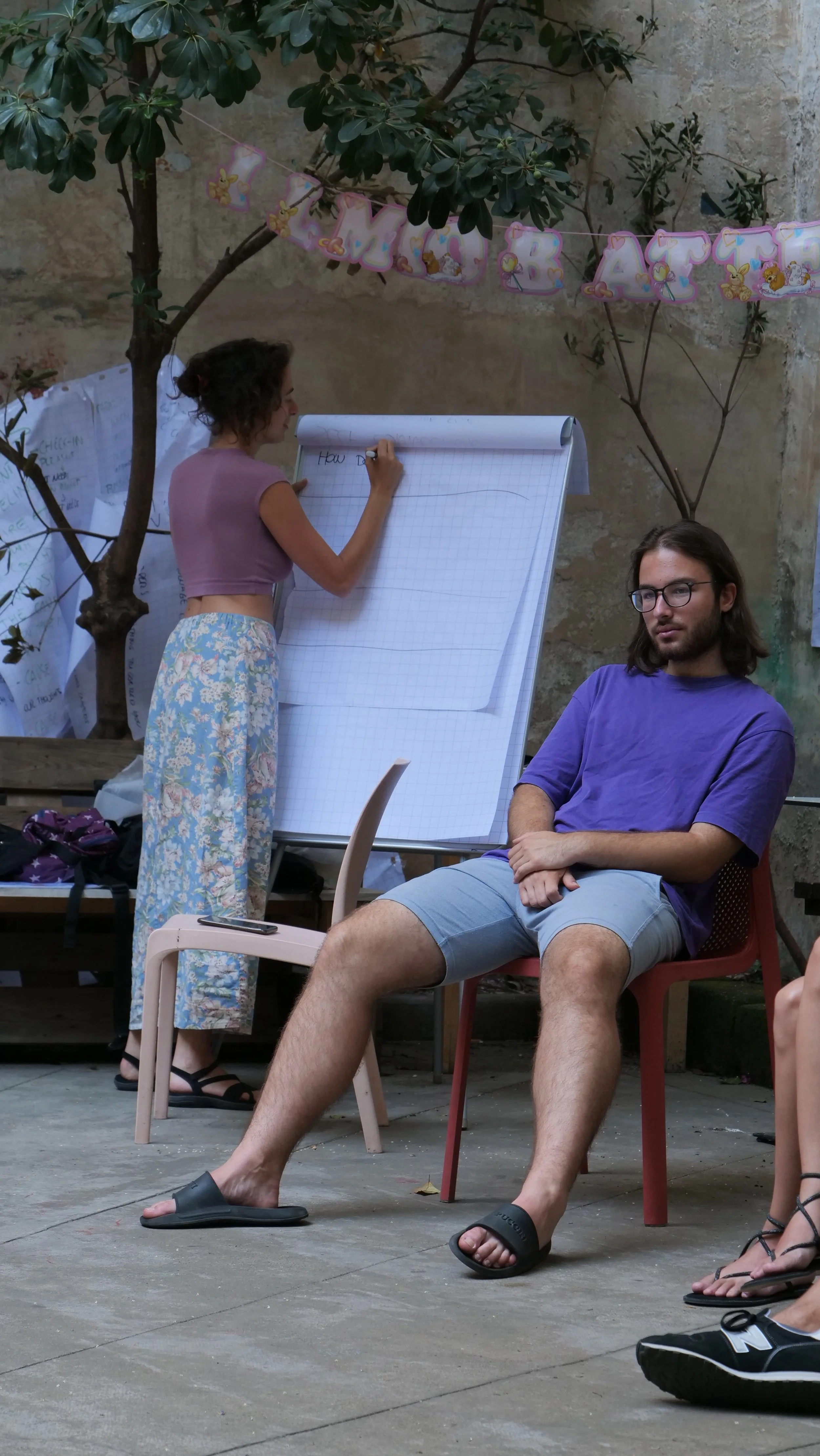 A woman and a man sitting outside in a shaded area, with the woman writing on a large notepad on an easel and the man sitting with one leg crossed over the other, wearing casual clothes and sandals. There are other people partially visible, a tree, and decorations hanging above.