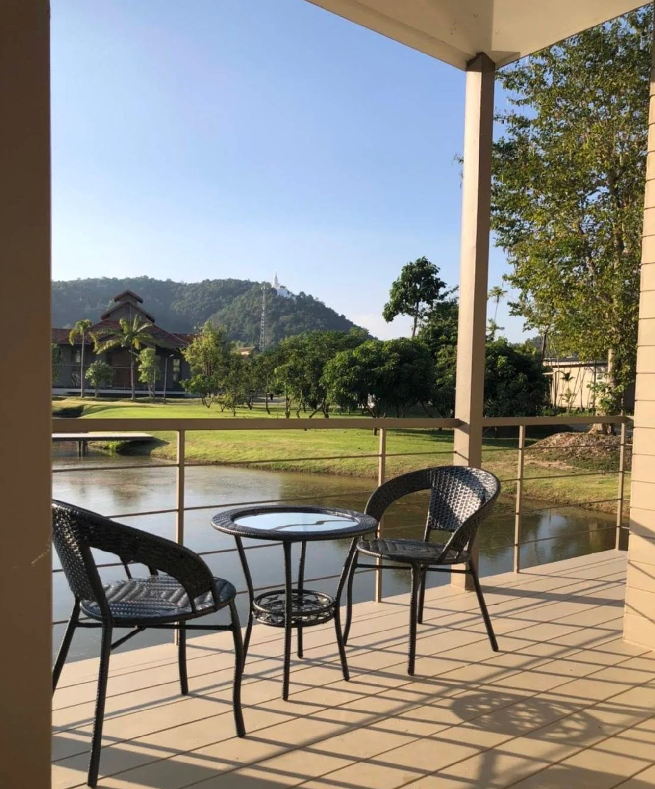 A balcony with a small round table and two black wicker chairs overlooking a pond with lush trees and a hill with a white structure in the background.