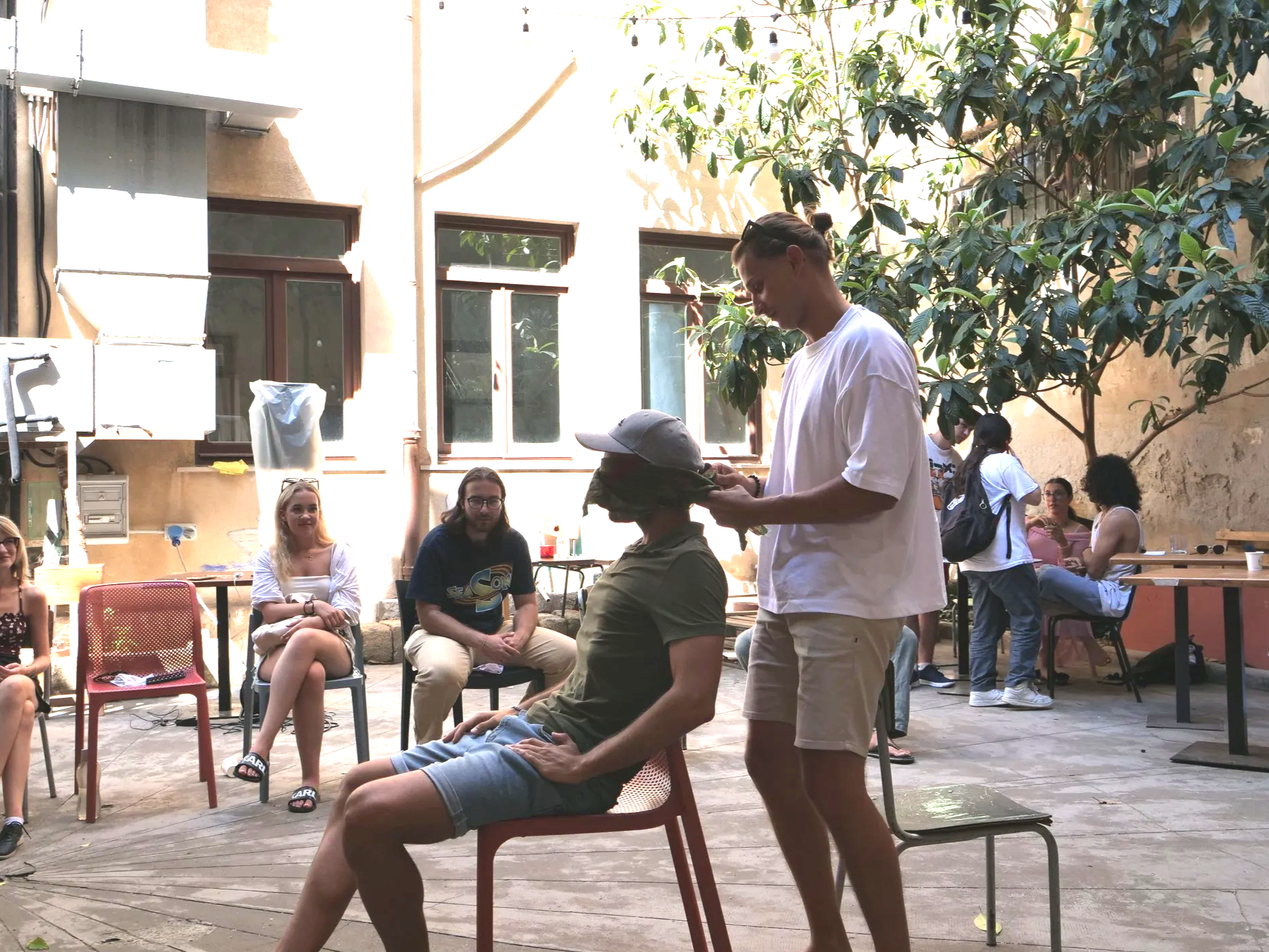 People sitting outdoors in a courtyard, with two individuals standing in the foreground. One person is seated, wearing a hat and green shirt, while the other is standing and holding a hat. Several others are seated and standing in the background, some with laptops, under a tree, with a building wall and windows behind them.