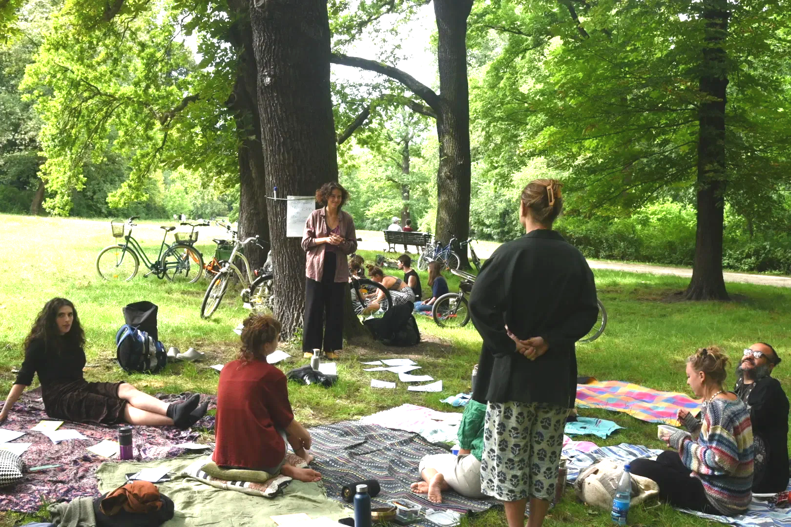 A group of people gathered outdoors in a park under trees, sitting or standing on blankets and surrounded by bicycles. Some are listening to a woman who is speaking or giving a presentation near a large tree.